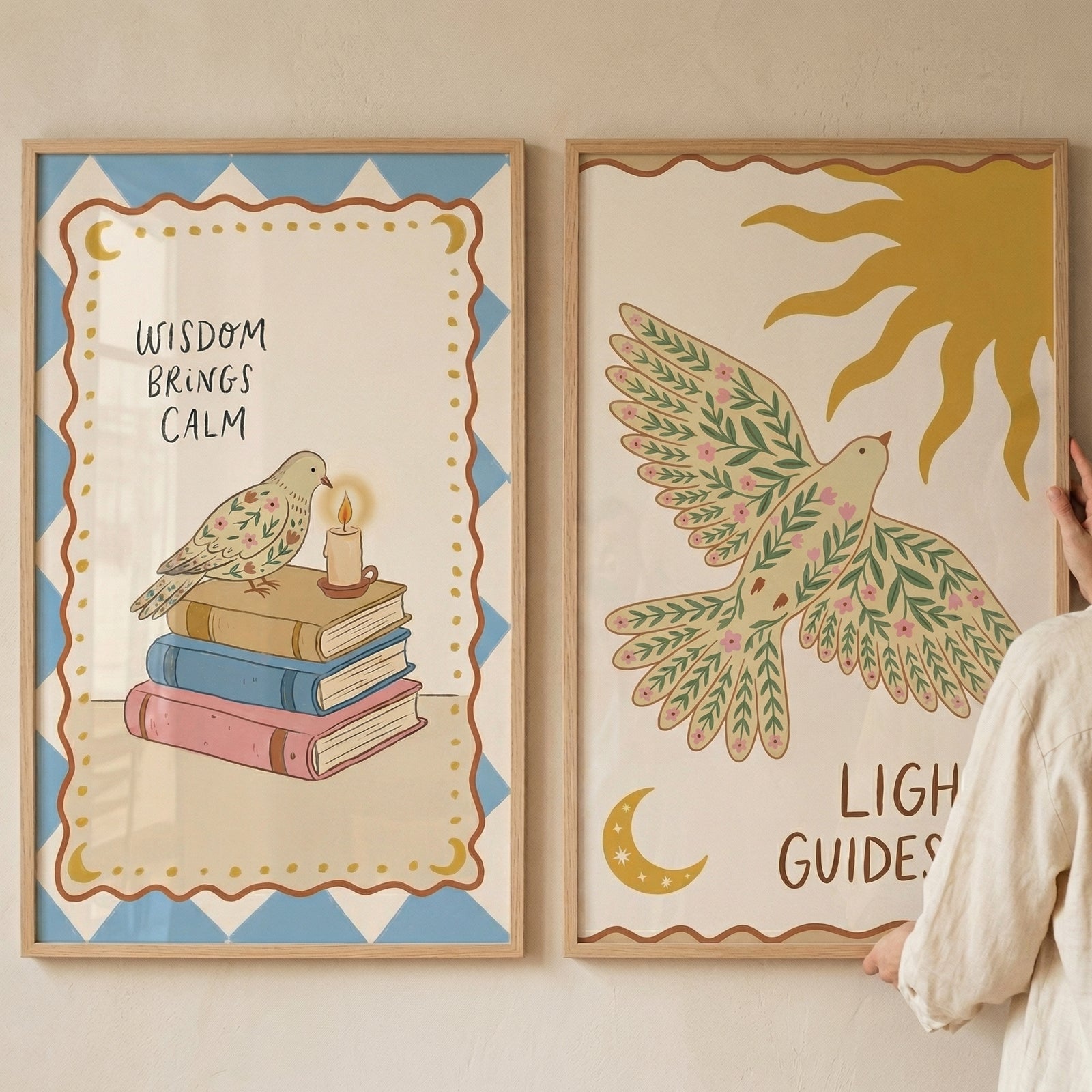 Two framed illustrations with a folk-art aesthetic hang side-by-side on a neutral wall. The left frame features a bird with floral wing patterns perched on a stack of three books—pink, blue, and yellow—next to a small, lit candle. 