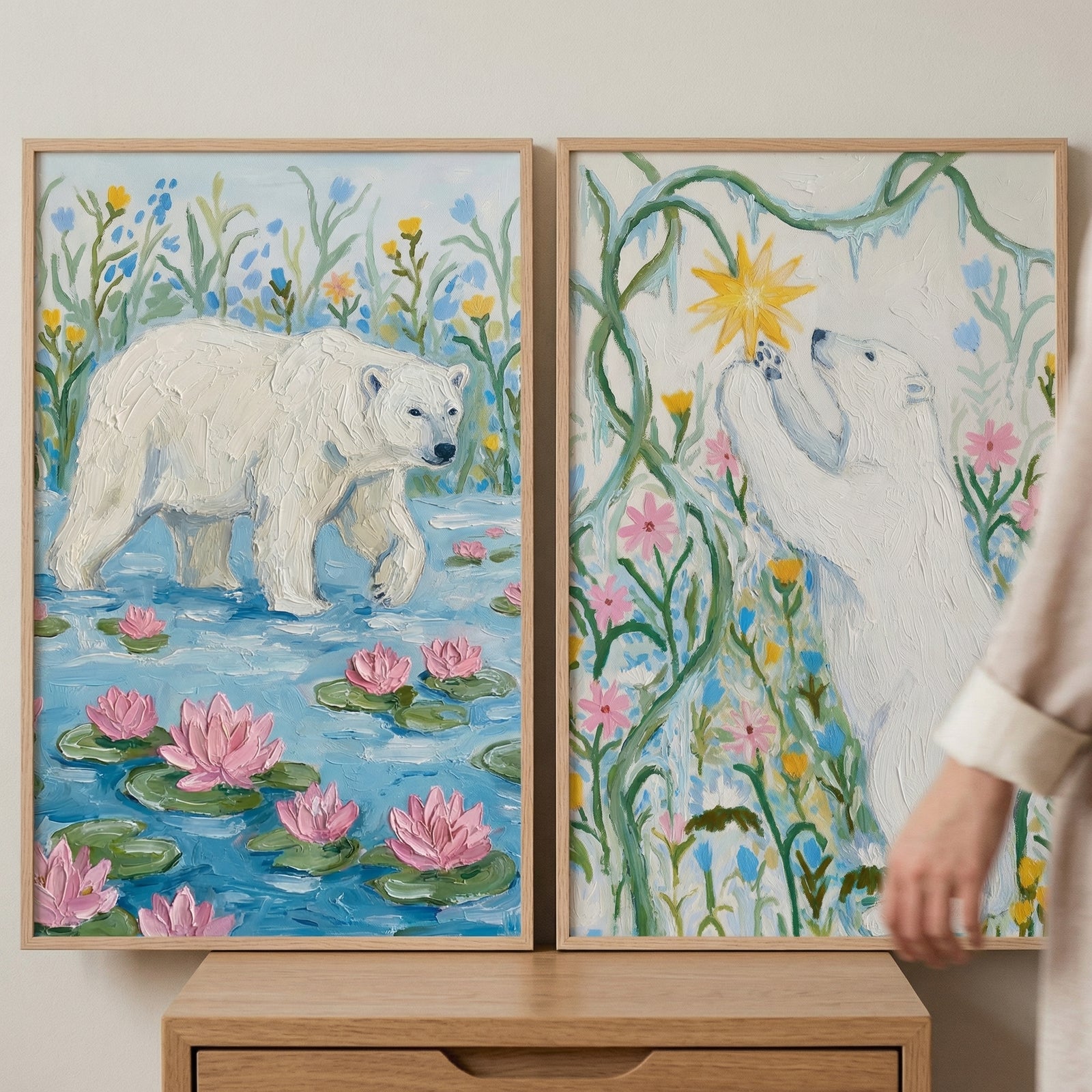 Two framed paintings of white polar bears displayed on a wooden surface, one showing a bear walking through blue water with pink lily pads and flowers, and the other reaching toward a glowing star among colorful blooms. A person’s hand enters the frame near the right artwork, suggesting the prints being arranged or styled.