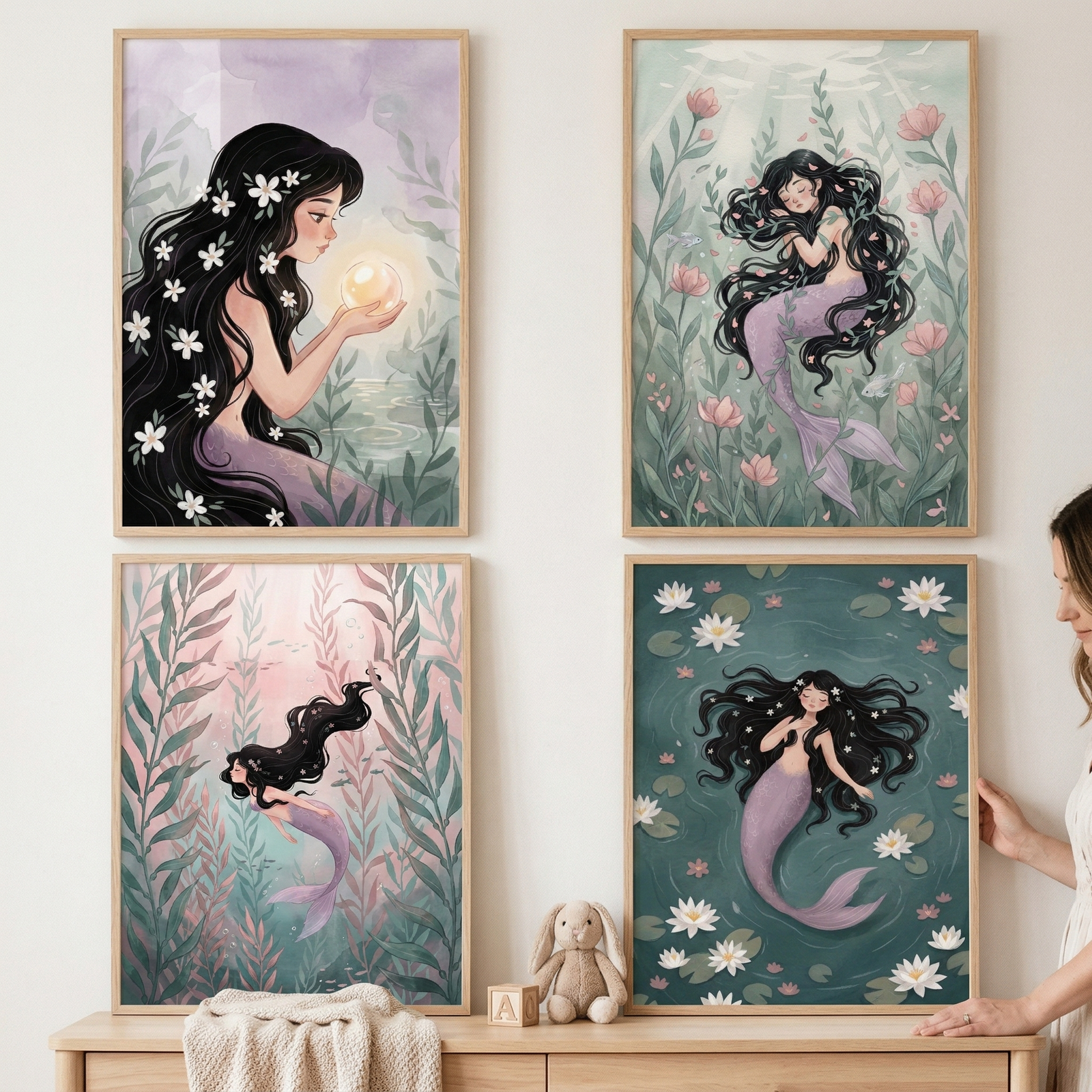 Four framed illustrations of a mermaid are displayed in a two-by-two grid on a light wall. The mermaid has long, dark hair with white flowers and a lavender tail. She is shown in various whimsical settings: holding a glowing orb, resting in an underwater garden, swimming through tall plants, and floating among water lilies. Below the light wood frames is a wooden dresser holding a small plush rabbit. A person's hand is visible on the right side, touching one of the frames.
