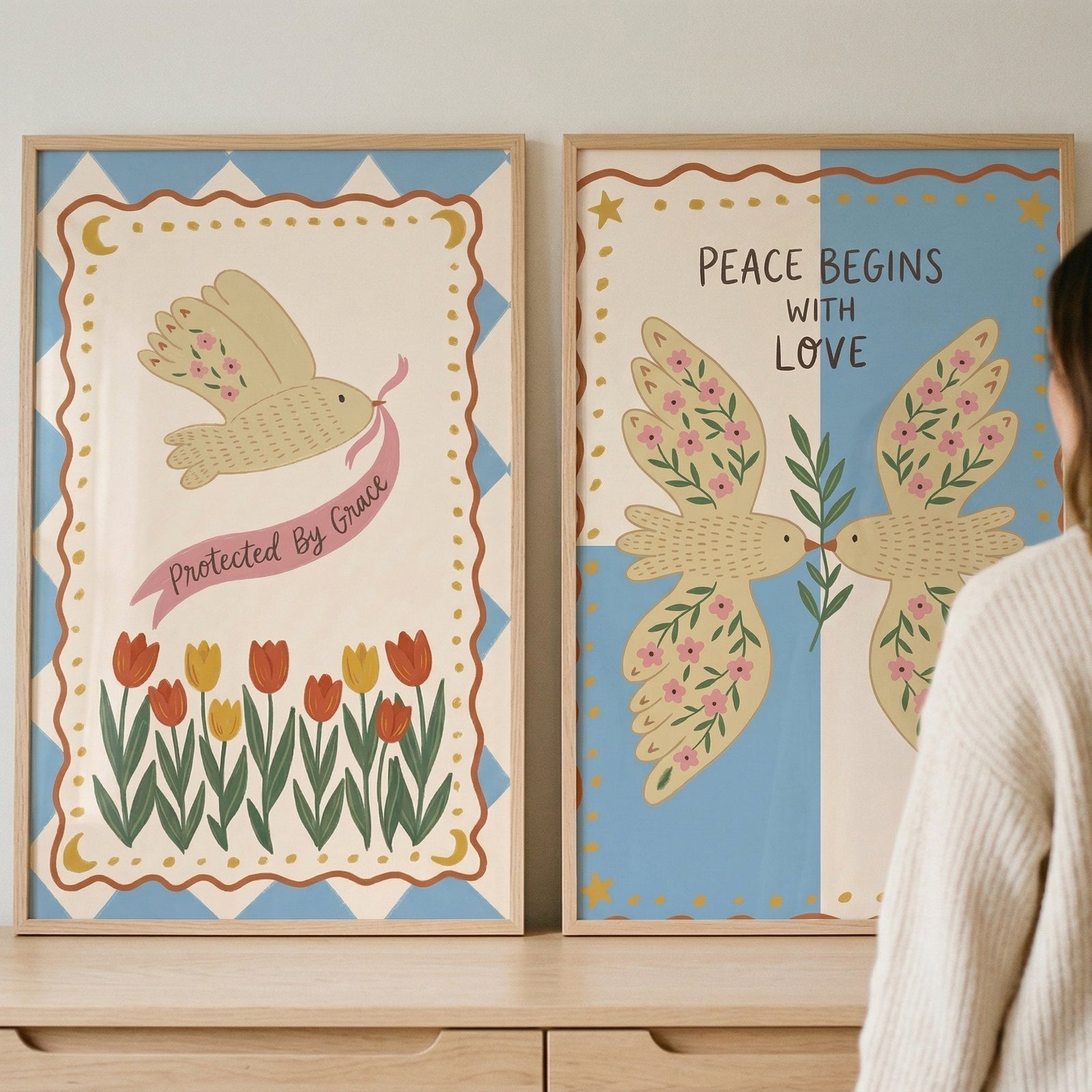 Two framed graphic art prints are displayed side-by-side on a light-colored wooden dresser. The print on the left features a stylized beige bird with pink floral patterns on its wings, flying and carrying a pink ribbon that reads "Protected By Grace." 