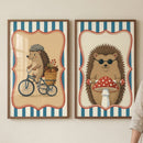 Two framed illustrations of anthropomorphic hedgehogs hang side-by-side on a light, neutral-toned wall. Both pieces feature a central character within a decorative, scalloped red frame, set against a background of vertical blue and white stripes. In the frame on the left, a hedgehog wearing a grey flat cap is depicted riding a blue tricycle. A small wicker basket on the back of the tricycle is filled with acorns and a sprig of red berries. The interior background behind the character is a light tan color.