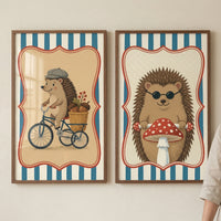 Two framed illustrations of anthropomorphic hedgehogs hang side-by-side on a light, neutral-toned wall. Both pieces feature a central character within a decorative, scalloped red frame, set against a background of vertical blue and white stripes. In the frame on the left, a hedgehog wearing a grey flat cap is depicted riding a blue tricycle. A small wicker basket on the back of the tricycle is filled with acorns and a sprig of red berries. The interior background behind the character is a light tan color.