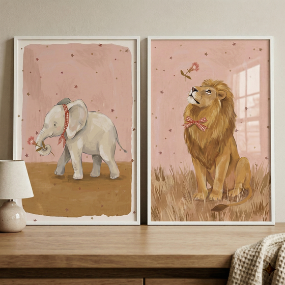 Two white-framed illustrations are displayed on a light wood surface. The left print shows a small elephant with a red and white checkered ribbon holding a pink flower. The right print features a lion with a matching bow tie gazing at a floating pink flower. Both have dusty pink backgrounds with tiny brown stars. 