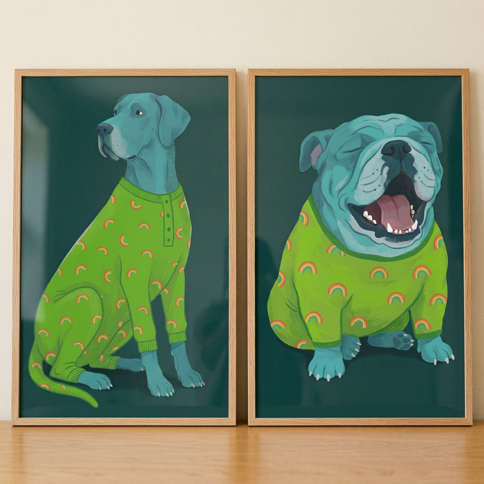 Two matching framed illustrations are displayed side-by-side on a light wood surface. Both pieces feature teal-colored dogs against a deep green background. On the left, a tall, slim dog is depicted in profile, sitting calmly and wearing a lime green onesie adorned with a pattern of small rainbows.