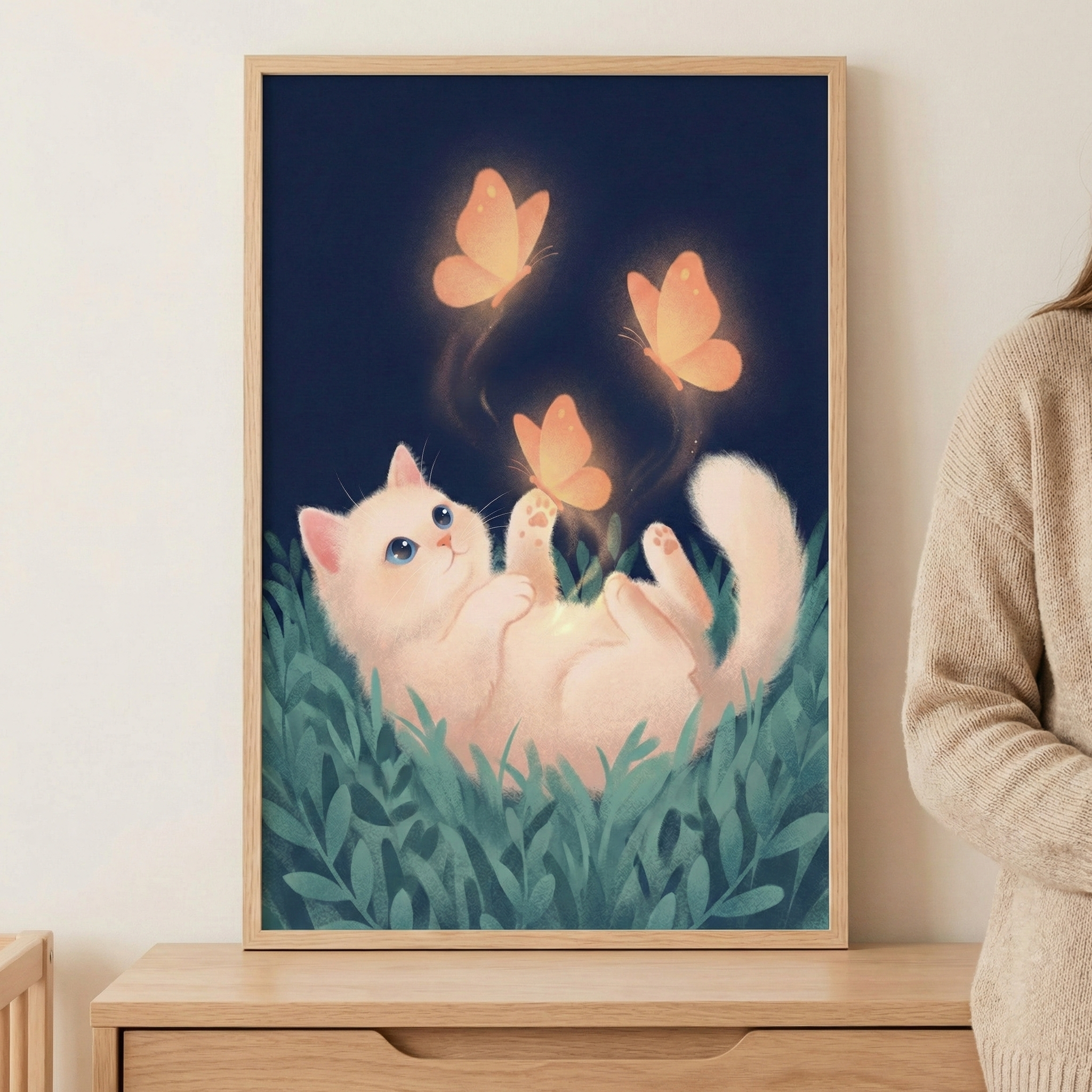 Framed wall art of a white kitten with blue eyes lying on its back in green grass, reaching up to touch three glowing orange butterflies against a deep navy night sky. The whimsical illustration adds a warm, magical feel to a child’s bedroom or nursery decor.