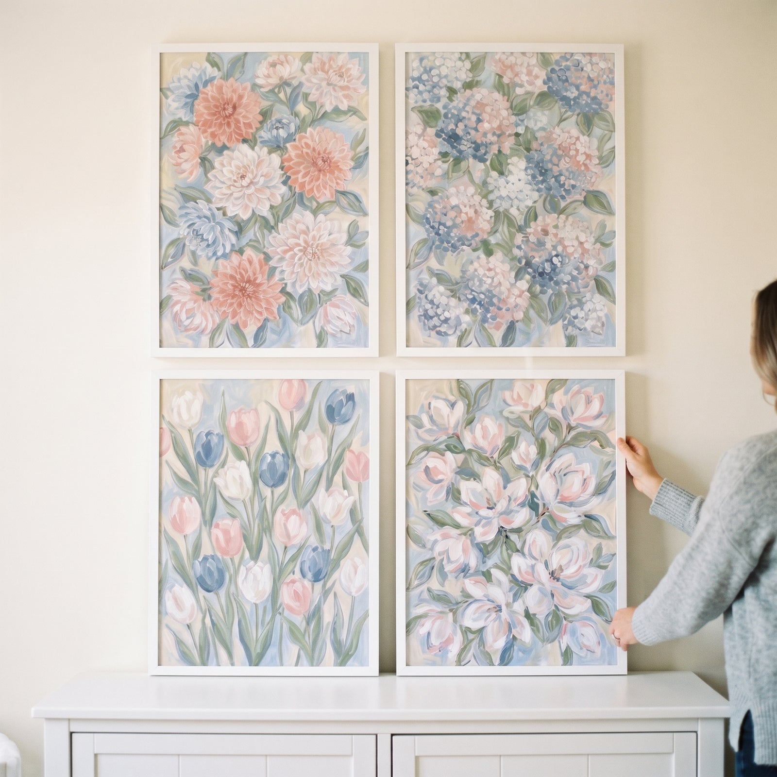 Hands adjust a whimsical pastel garden floral wall art set of 4 displayed in white frames on a light wall.