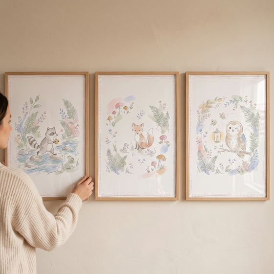 A person hangs a whimsical pastel woodland animals wall art set of 3 featuring a raccoon, fox, and owl in wooden frames.