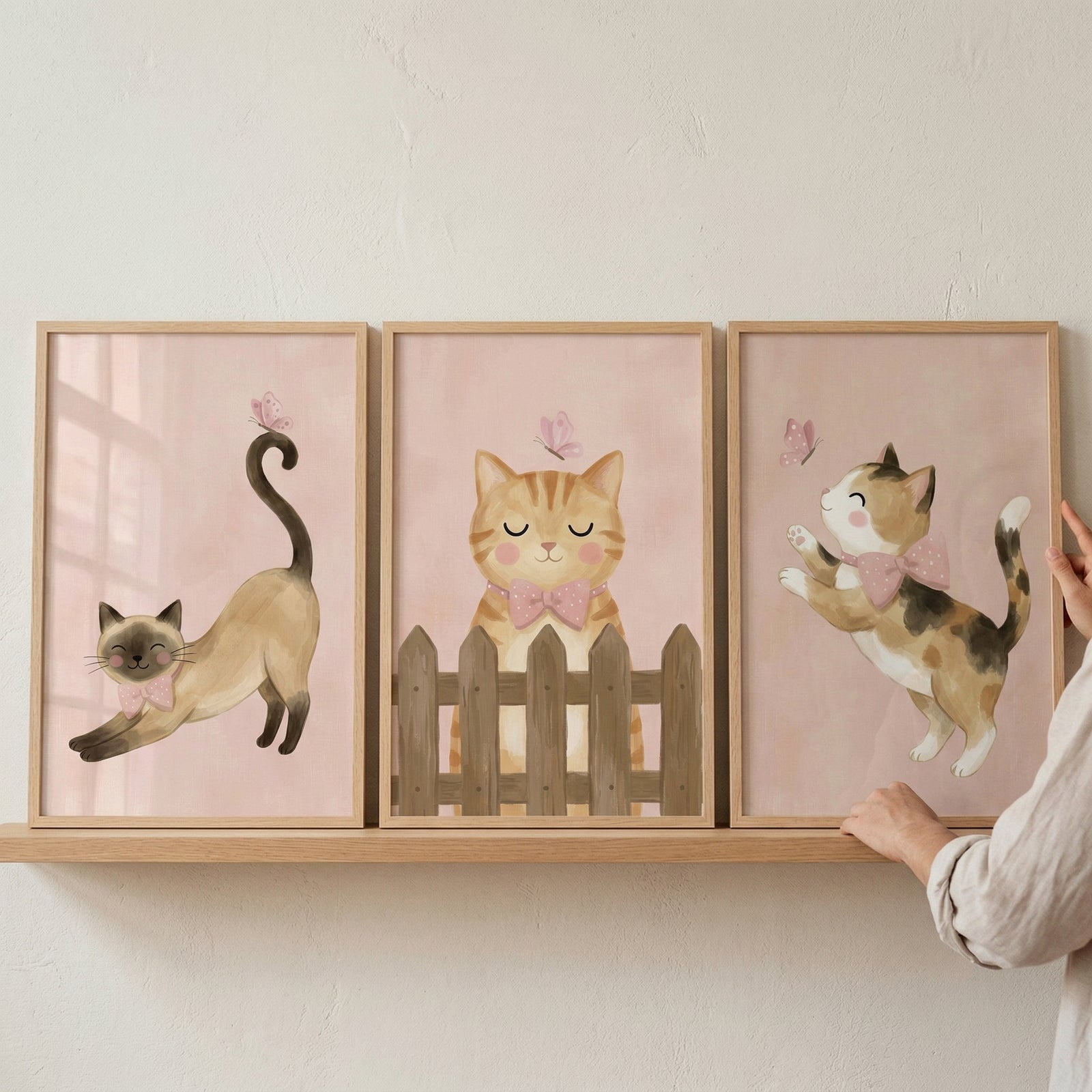 Three framed watercolor-style illustrations of playful cats are displayed on a light wooden shelf against a plain wall. Each piece is housed in a thin, light-wood frame and features a soft pink background.

