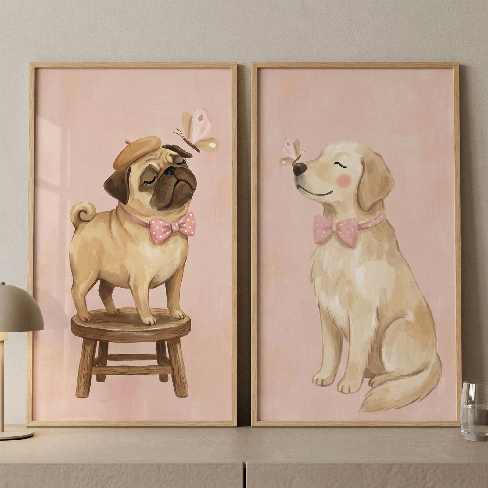 Two side-by-side vertical illustrations in light wood frames are displayed on a neutral surface against a soft-toned wall. Both pieces feature a gentle, whimsical style with soft pink backgrounds. 
