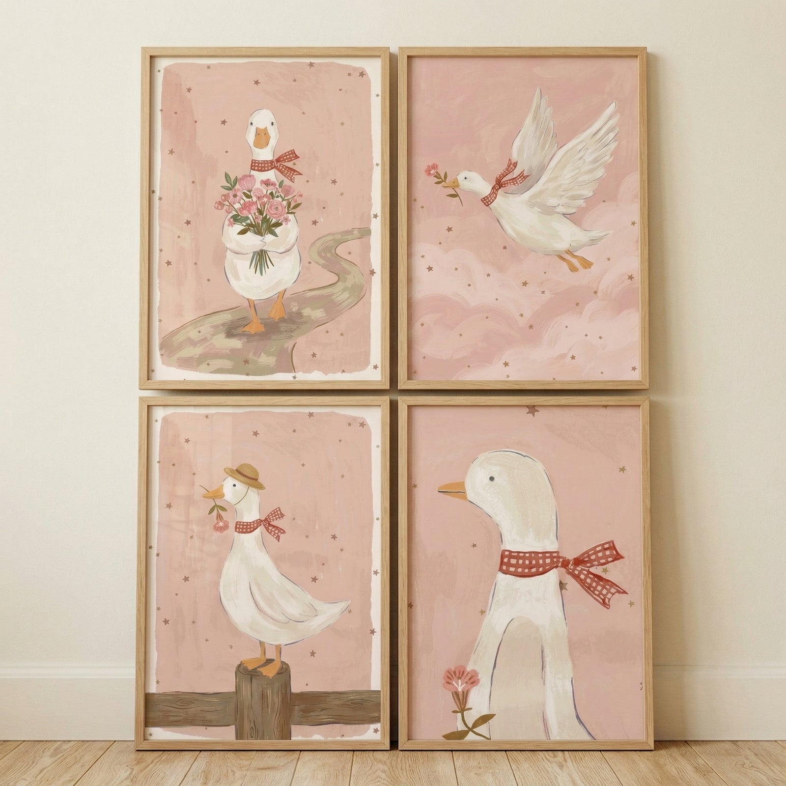 Four framed nursery prints arranged in a grid show soft pink illustrations of ducks, including one holding a bouquet of flowers, one flying with a flower in its beak, one wearing a hat while perched on a post, and one close up with a red scarf. The coordinated artwork creates a sweet and whimsical focal point against a neutral wall.