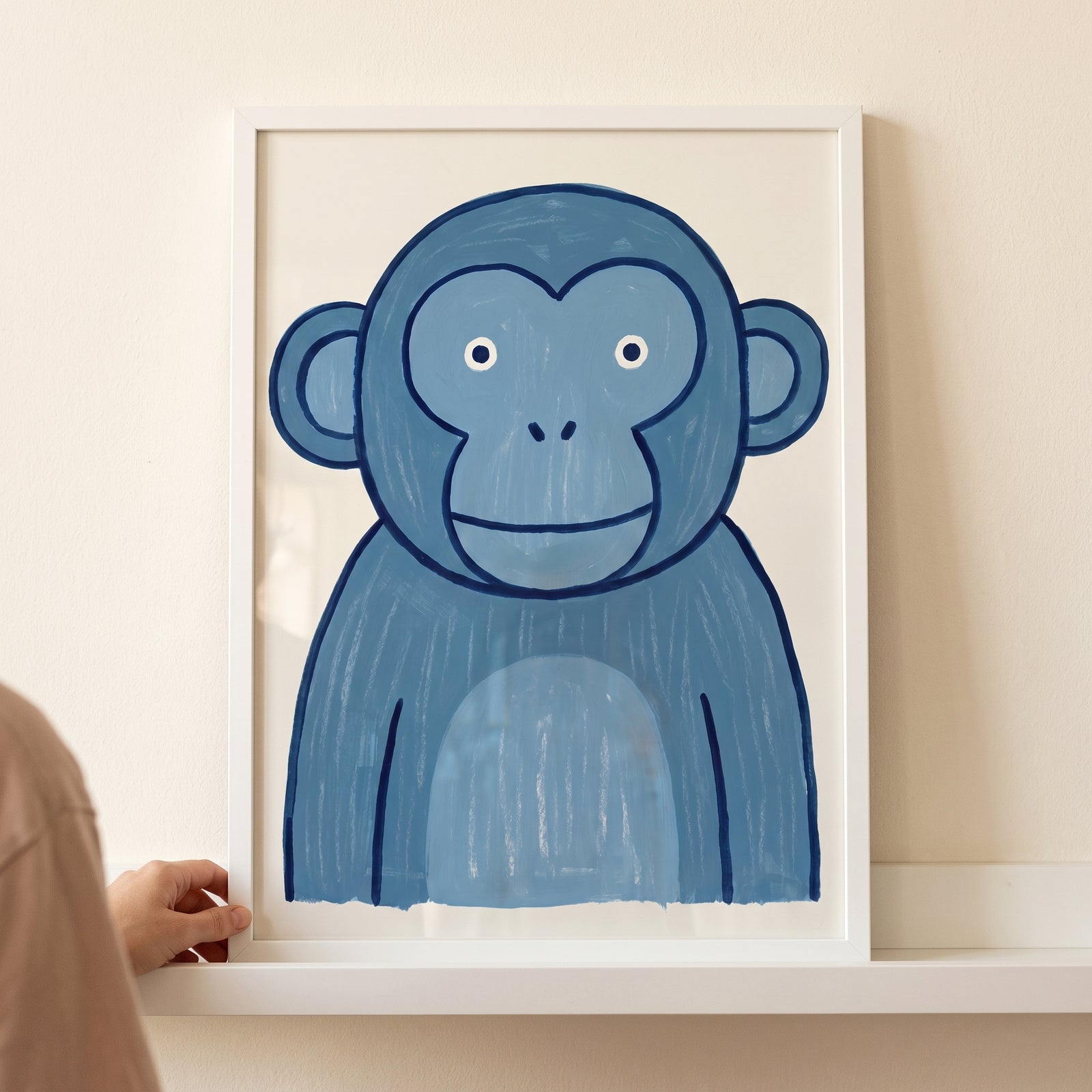Whimsical teal blue monkey wall art in a white frame held by a hand on a shelf.