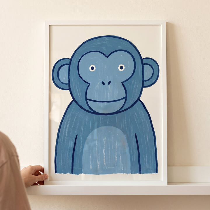 Whimsical teal blue monkey wall art in a white frame held by a hand on a shelf.