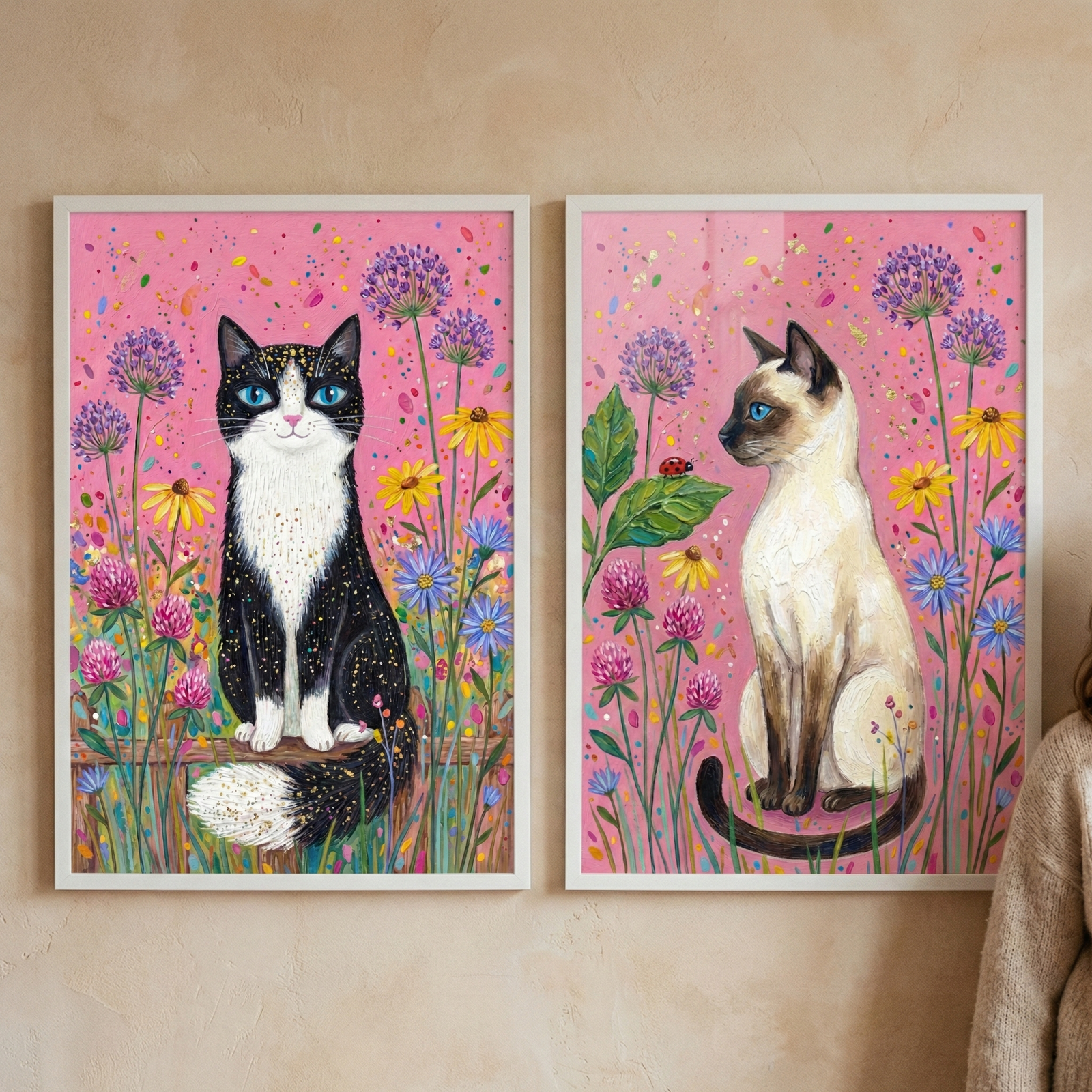 Two framed vertical illustrations of cats are displayed side-by-side. On the left, a black and white tuxedo cat with starry-textured fur and bright blue eyes faces forward in a garden of purple, yellow, and blue flowers against a pink sky. On the right, a light-colored cat with dark brown markings and blue eyes sits in profile among similar flora. The whimsical style features rich textures and a vibrant color palette, both set within simple white frames.