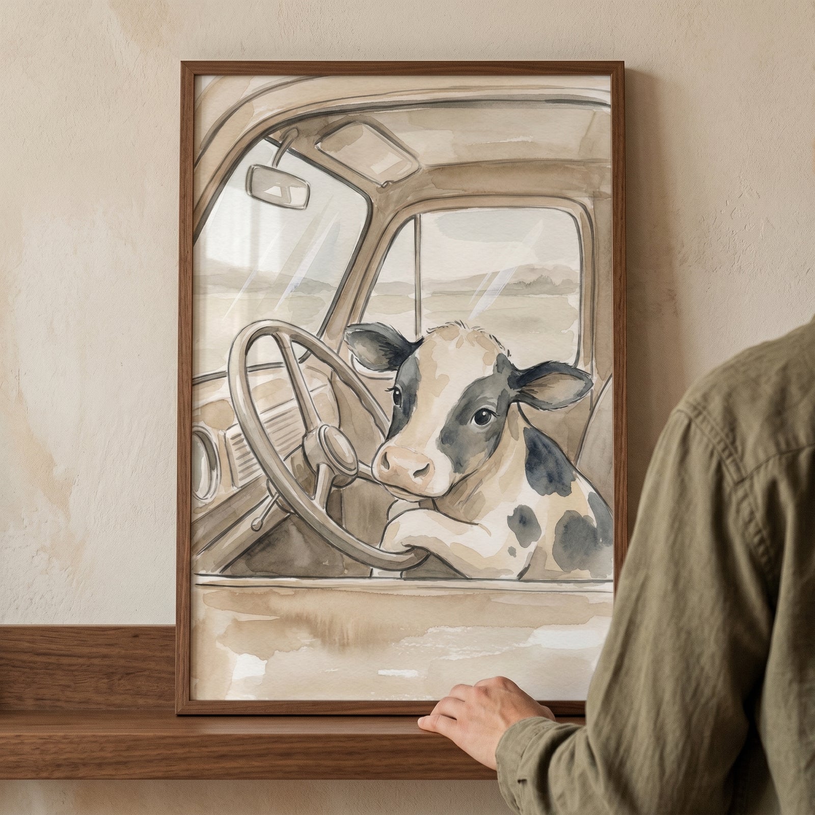 Framed whimsical watercolor driving cow wall art depicts a cow at the wheel of a vintage truck on a wooden shelf.