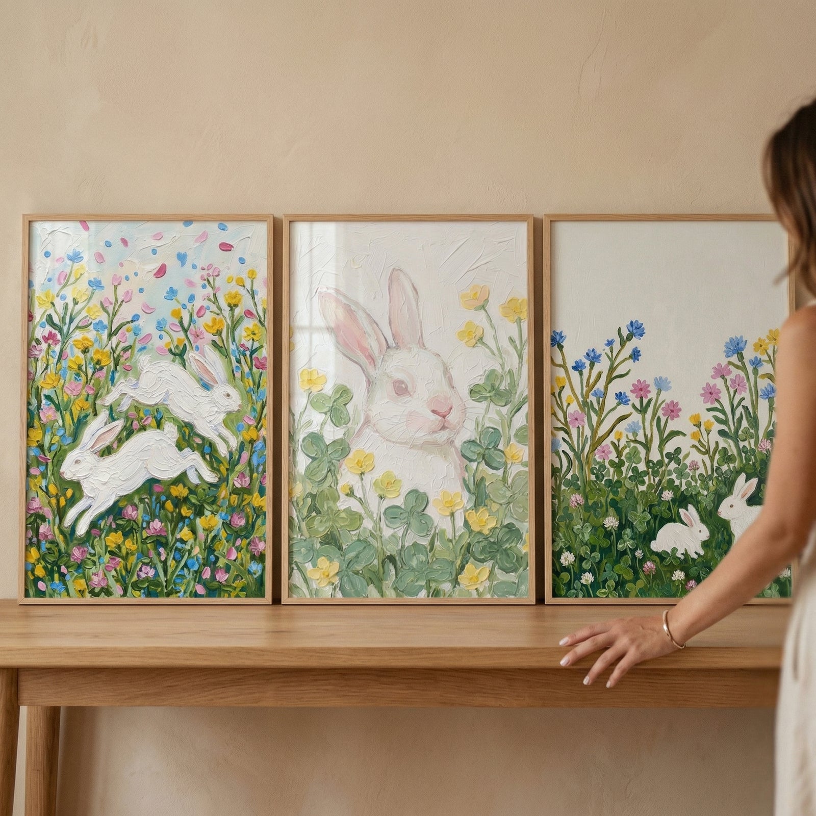 Three framed paintings of white rabbits are displayed on a wooden table against a neutral wall, each surrounded by colorful flowers and greenery. The left shows two rabbits leaping through a meadow of pink, yellow, and blue blooms, the center features a close up of a rabbit nestled among clover and small yellow flowers, and the right shows a rabbit sitting in a field of tall wildflowers while a person’s hand rests on the table nearby.