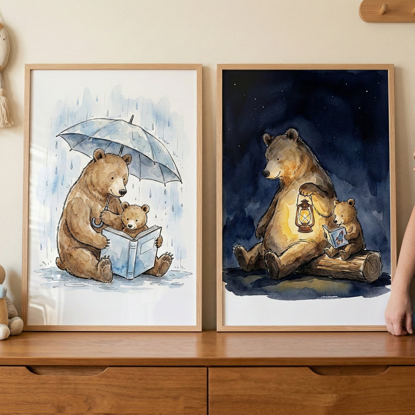Two framed art pieces are displayed side-by-side on a wooden dresser. Both feature a large brown bear and a small cub in a soft watercolor style. In the frame on the left, the two bears sit together in the rain. The adult bear holds a large blue umbrella over them while holding an open book for the cub. The background is a pale, rainy sky. The frame on the right shows the pair at night, sitting on a fallen log. The adult bear holds a glowing lantern, illuminating the scene as the cub looks at a small book.