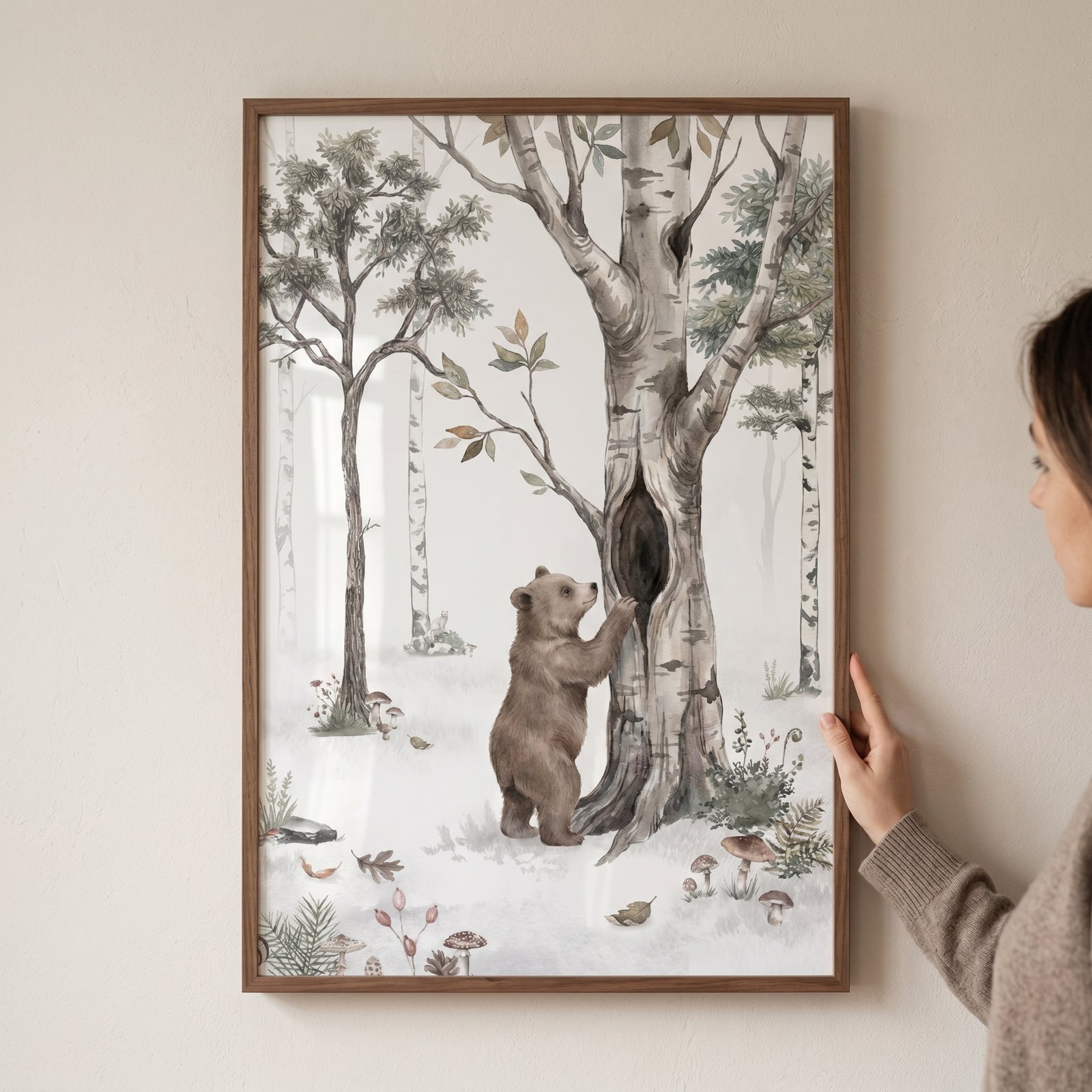 A hand holds whimsical woodland bear wall art featuring a bear cub by a hollow tree in a soft, illustrated forest.