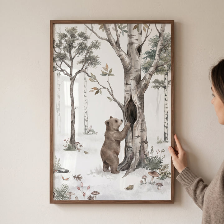 A hand holds whimsical woodland bear wall art featuring a bear cub by a hollow tree in a soft, illustrated forest.