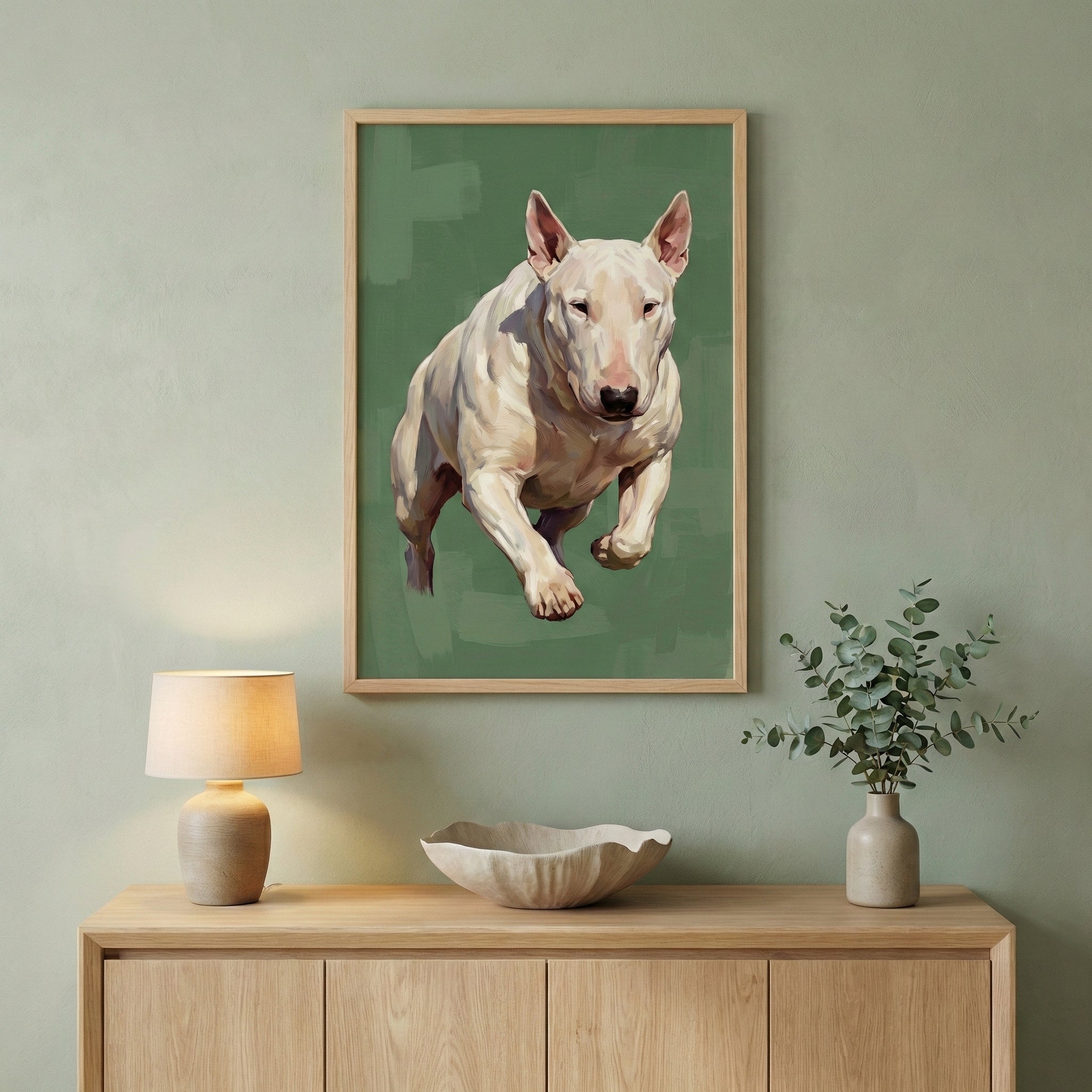 Framed painting of a white Bull Terrier mid-leap, displayed in a serene, modern interior. The dog is captured in a dynamic pose against a muted green background, with painterly brushstrokes emphasizing its strong, muscular form. The artwork hangs above a light wood sideboard adorned with minimal, natural decor— a ceramic lamp, a sculpted bowl, and a vase with eucalyptus branches—creating a warm and balanced aesthetic.