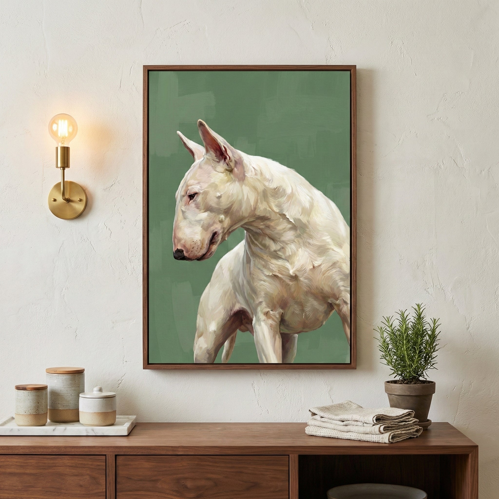 A cozy, minimalist interior featuring a framed painting of a white Bull Terrier on a green background. The dog is depicted in a realistic, painterly style, looking to the side with a calm, muscular stance. The artwork is hung above a wooden console with natural-toned decor including ceramic containers, folded linens, and a potted herb plant, all softly lit by a brass wall sconce.