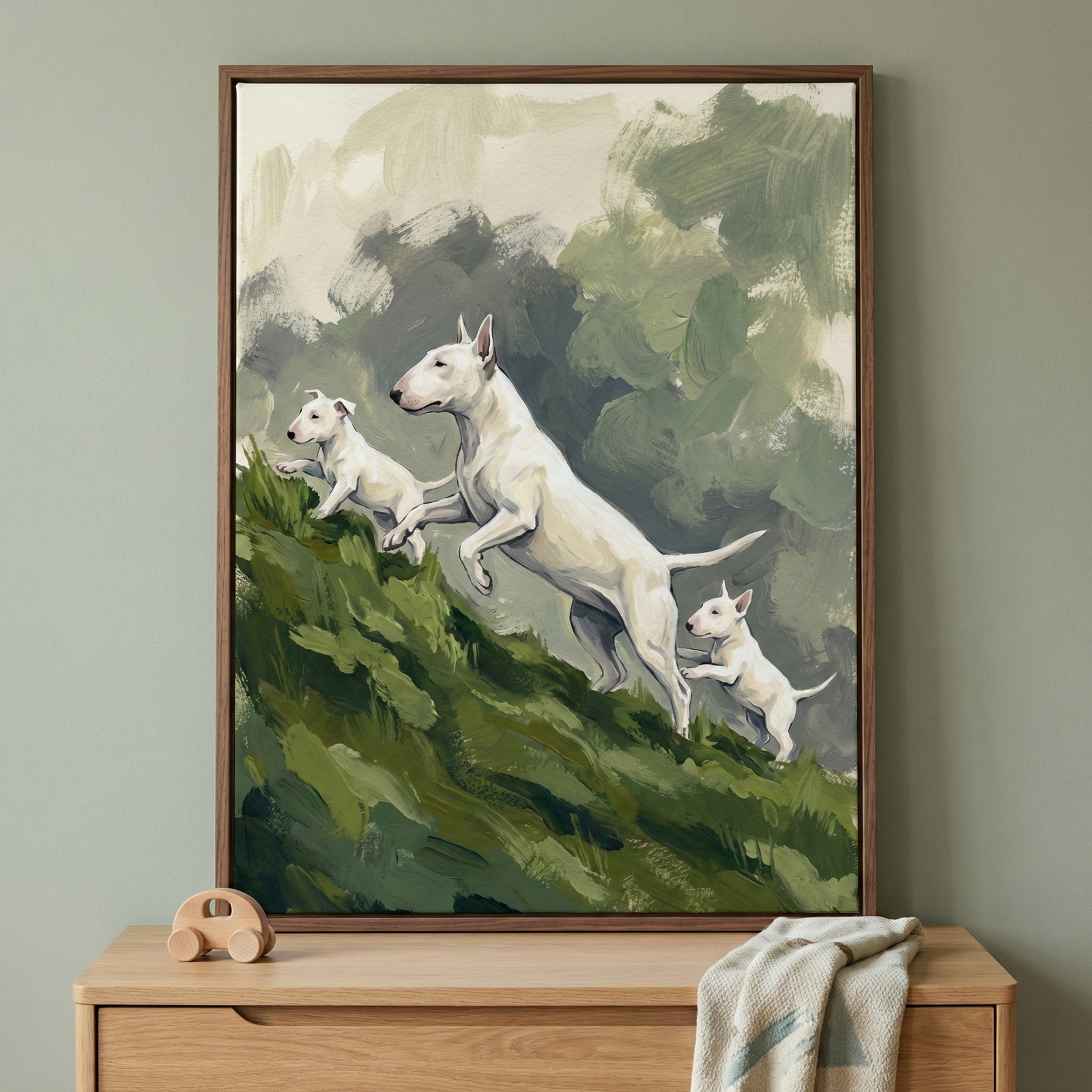 A framed piece of artwork hangs on a sage green wall above a light wood dresser. The artwork depicts an adult white dog with a distinctive egg-shaped head leaping up a lush green slope, followed by two smaller puppies. The scene is created with broad, visible brushstrokes and a palette of earthy greens and soft greys. On the wooden dresser below the frame, a small toy car and a folded cloth rest, completing a peaceful interior scene.