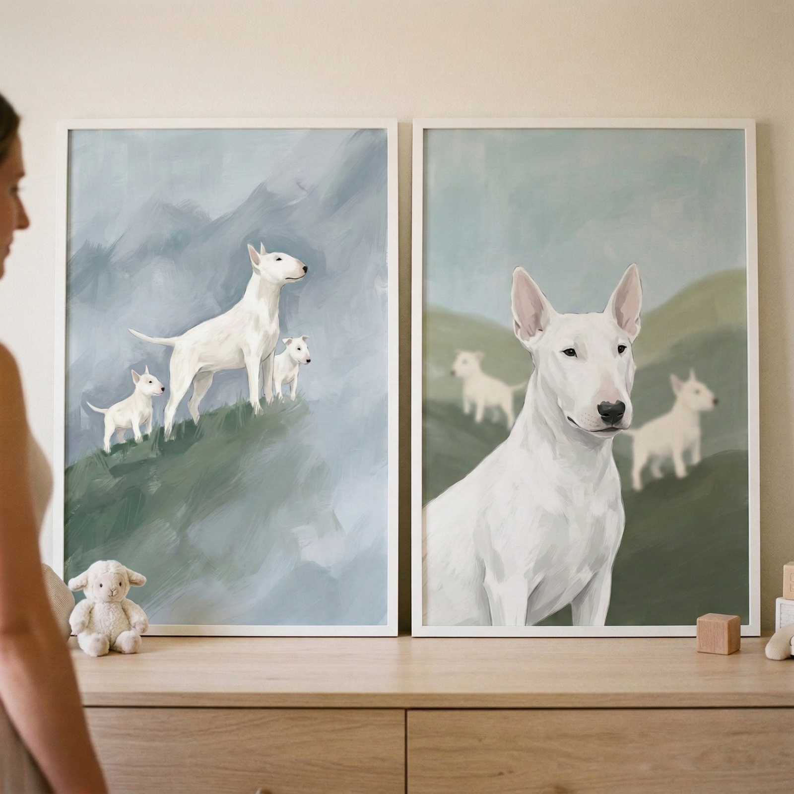 Two white-framed vertical art prints sit on a light wood dresser. The print on the left shows a large white bull terrier and two smaller puppies standing on a green grassy hill with a misty blue-grey mountain landscape behind them. The print on the right features a close-up of a white bull terrier's face and chest, with two more dogs in a soft-focus background of green hills. A woman is partially visible on the left side of the frame, looking toward the art. On the dresser in front of the prints are a small