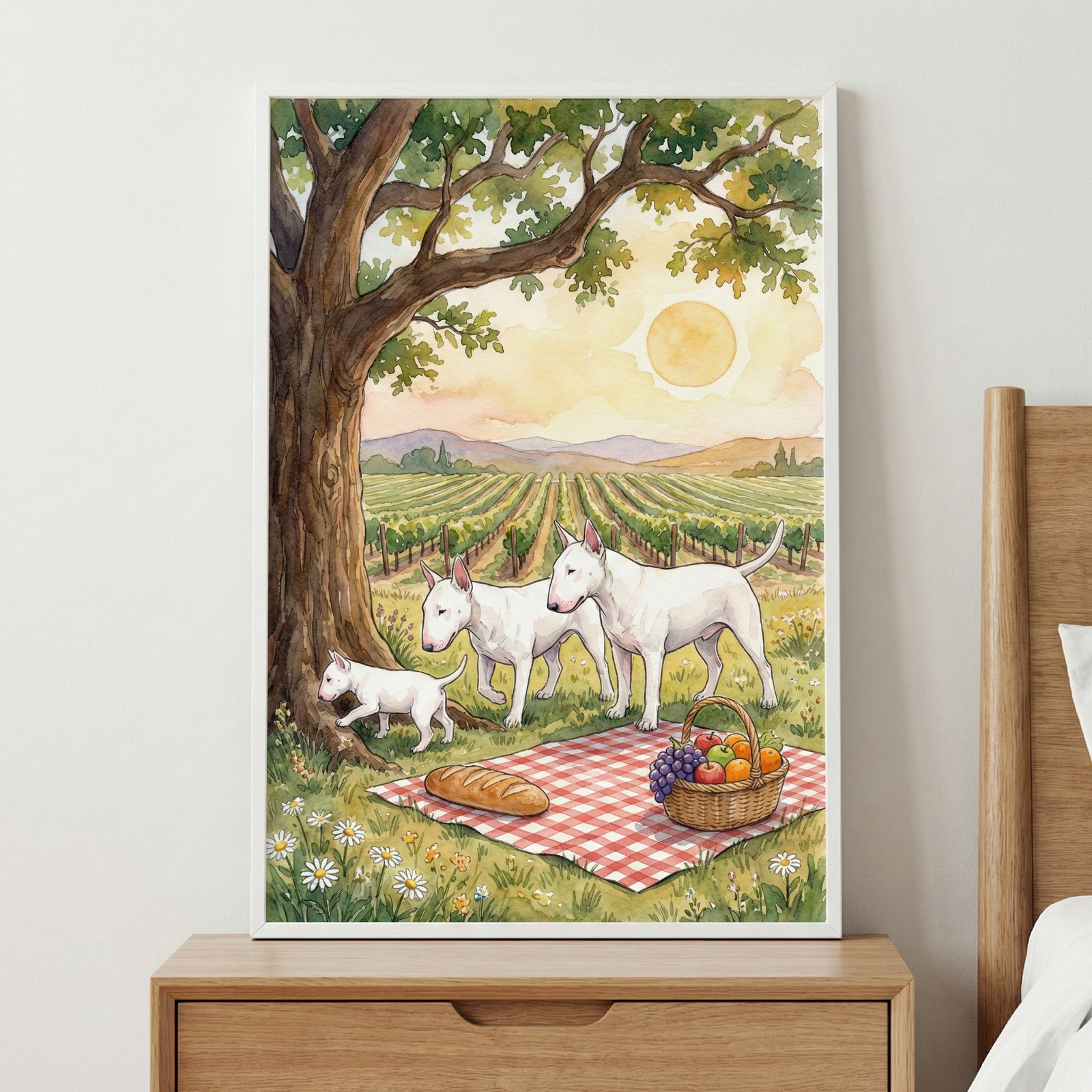 A framed watercolor illustration shows three white bull terriers—two adults and a small puppy—near a picnic in a vineyard. On a red-and-white checkered blanket lies a loaf of bread and a basket of fruit. The dogs stand under the shade of a large, leafy tree. In the distance, rows of grapevines stretch toward rolling hills under a hazy, warm sky with a prominent golden sun. The artwork is displayed in a white frame on a wall above a wooden bedside table.