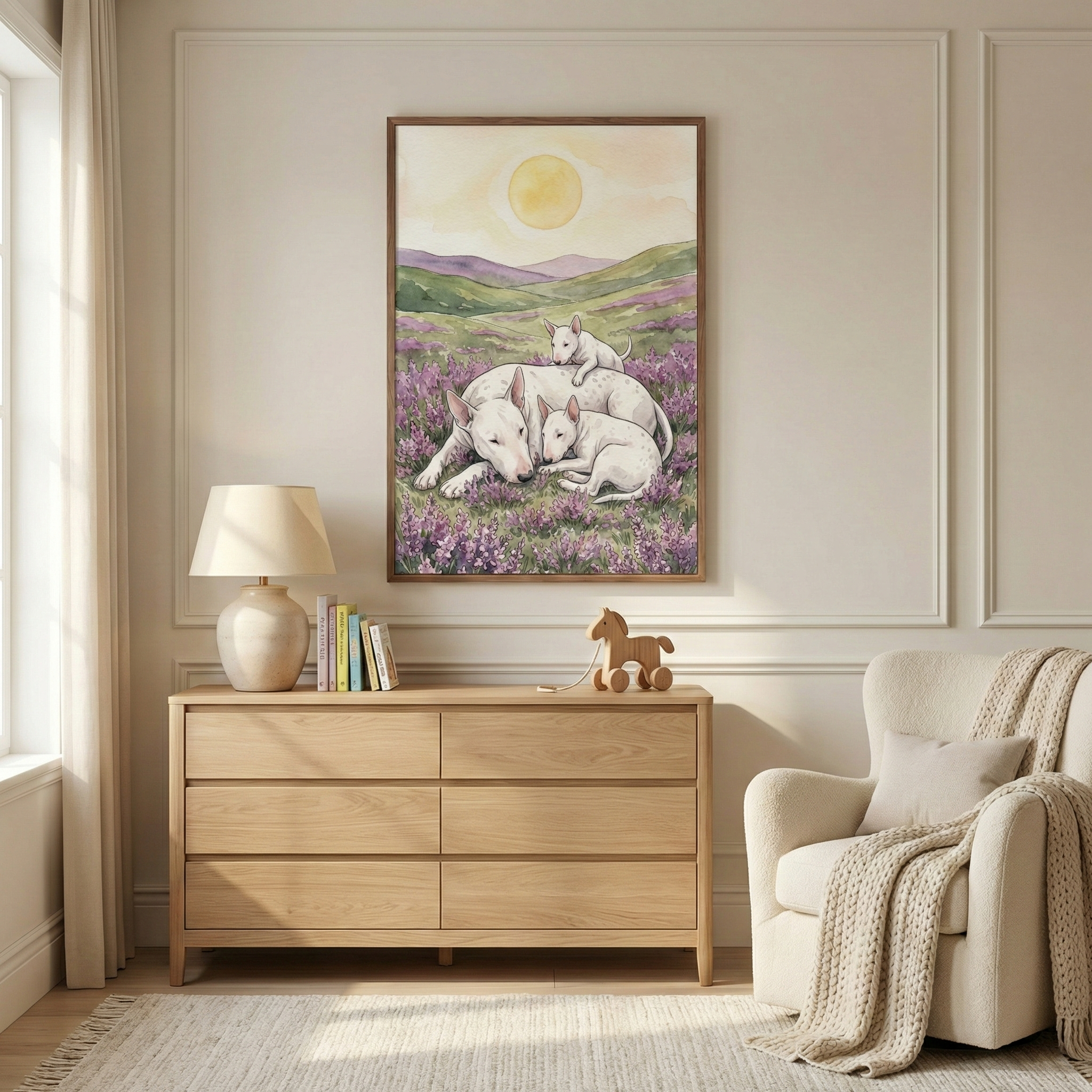 A bright, minimalist room features a large vertical framed illustration of a mother white dog with distinct facial features and her two puppies in a field of purple flowers. The dogs are resting under a large yellow sun and rolling green hills. The artwork hangs above a light wood dresser holding a lamp, books, and a wooden toy horse. Beside the dresser is a cozy cream-colored armchair with a textured throw blanket. The room has off-white walls and a light-colored rug.