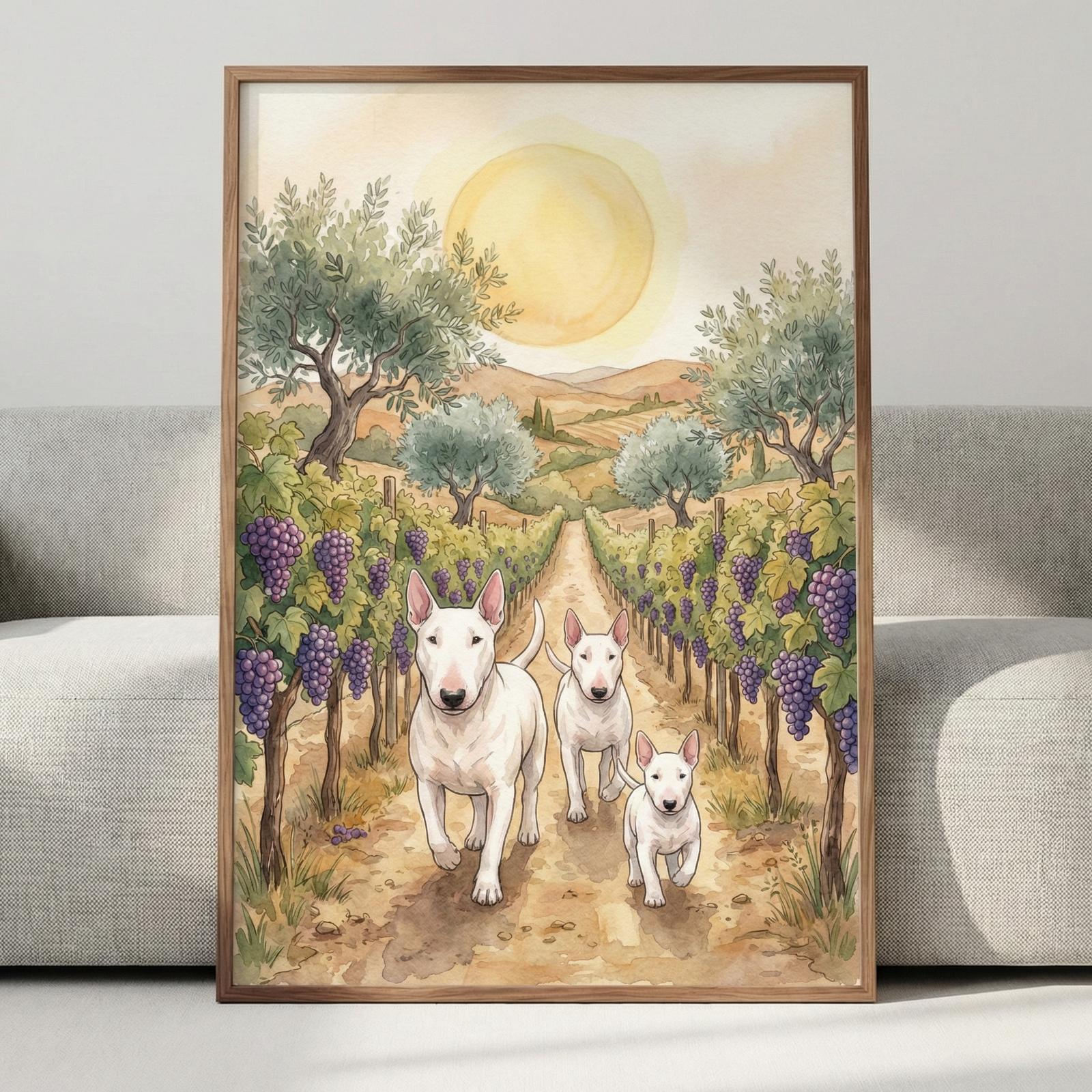 A framed watercolor-style illustration leans against a gray sofa. The artwork features three white bull terriers of varying sizes walking down a dirt path in a sun-drenched vineyard. Purple grapes hang from green vines on either side of the path. In the background, rolling hills and a few trees are visible under a large, soft yellow sun in a pale sky. The overall aesthetic is calm and illustrative with gentle color washes.