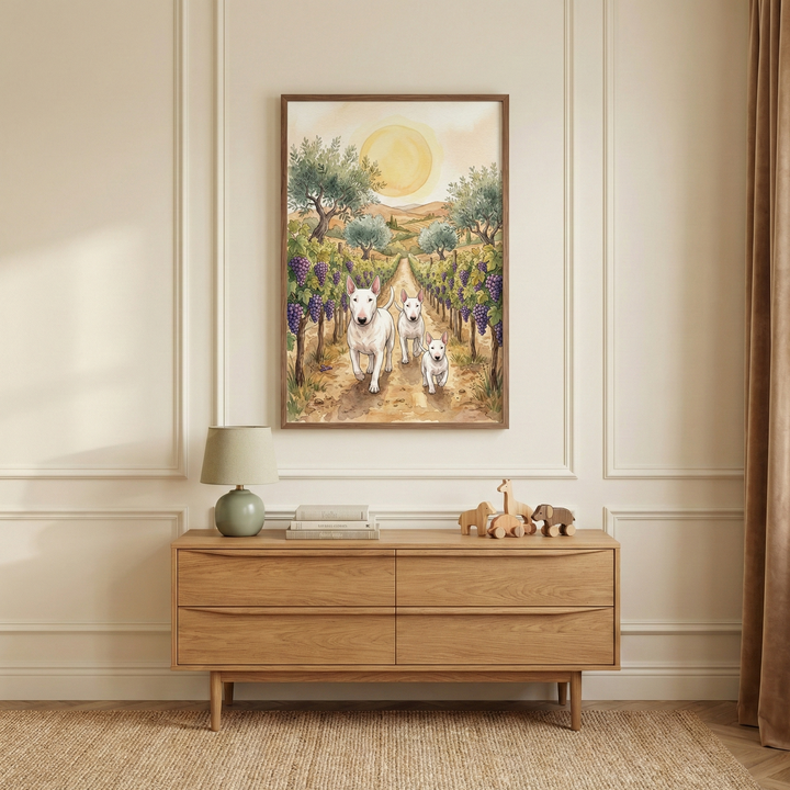 A framed watercolor-style illustration hangs on a neutral-toned wall with rectangular molding. The art depicts three white terriers walking down a dirt path through a lush vineyard. Purple grapes cluster on the vines, and the background features rolling hills and olive trees under a large, soft sun. Below the art is a light-wood four-drawer dresser. On the dresser sit a small green lamp, a stack of books, and several wooden animal figures. A natural-fiber rug is on the floor.