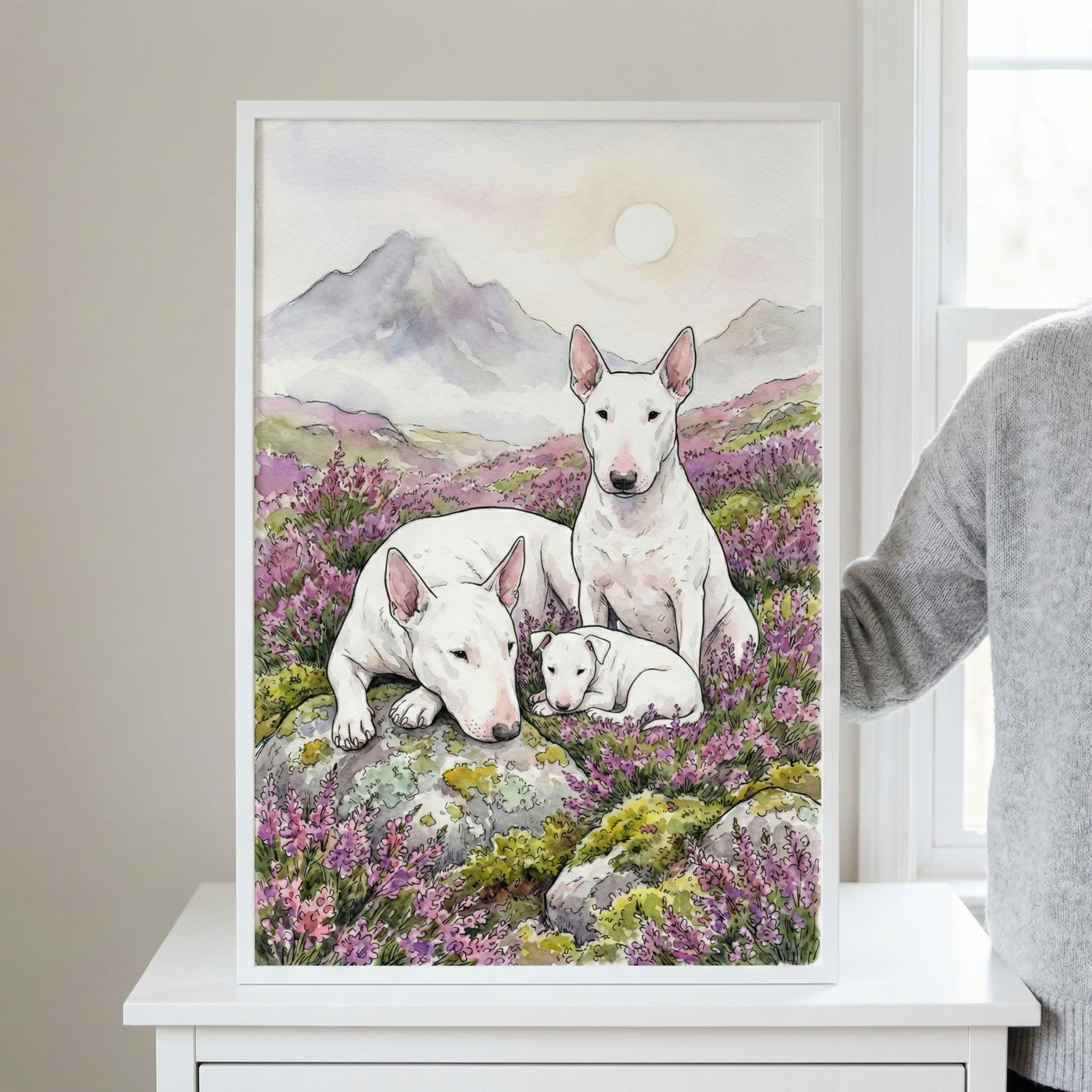 A white-framed artwork displays three white dogs with distinctive egg-shaped heads resting in a field of vibrant purple heather. A large adult dog sits upright, another lies peacefully across a mossy rock, and a small puppy is nestled between them. The scene is set against a backdrop of soft, misty mountains under a pale, hazy sky with a large glowing sun. The illustration uses delicate ink lines and soft watercolor washes. 