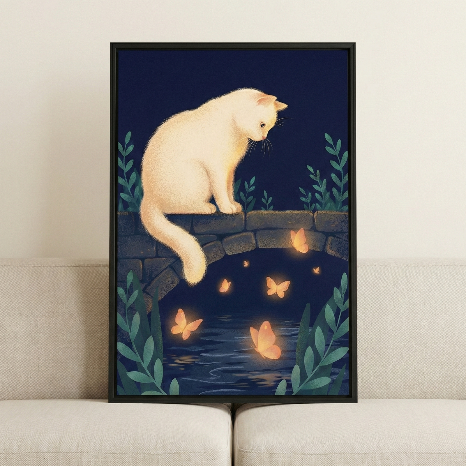 A framed illustration of a fluffy white cat sitting on a low stone bridge during the night. The cat is looking down at several glowing orange butterflies that are fluttering over the dark water visible beneath the bridge's arch. Stylized green plants grow on either side of the scene against a deep blue background. The framed artwork is propped up on a light-colored sofa against a neutral wall.