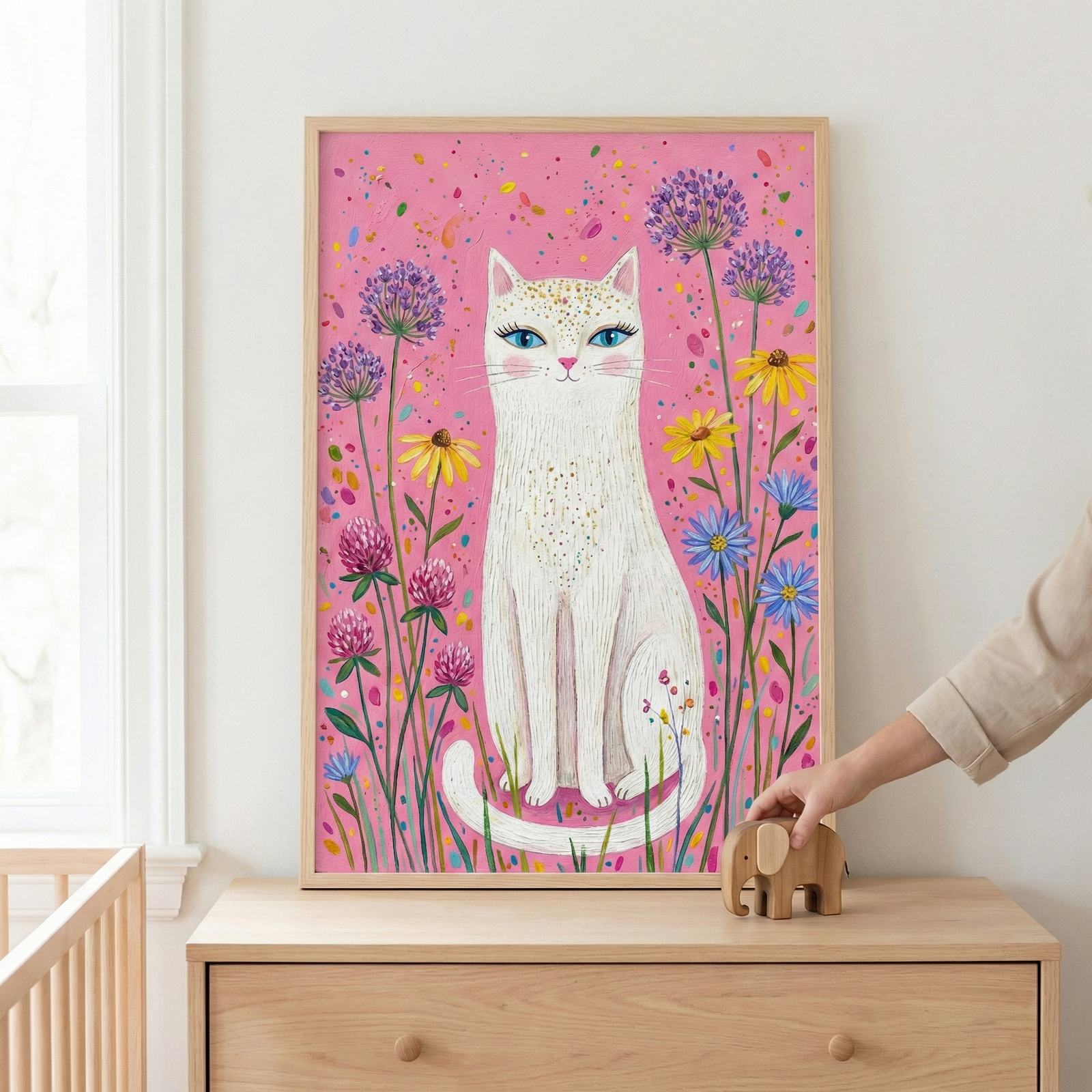 This framed illustration features a stylized white cat with striking blue eyes and gold-speckled fur. It is centered against a bright pink background filled with colorful splatters and an array of whimsical flowers, such as purple globes, yellow daisies, and blue blossoms. The artwork sits in a light wood frame on a matching dresser. Beside the frame, a person's hand reaches for a small wooden elephant toy. The scene is set in a light-filled room with a wooden crib partially visible.