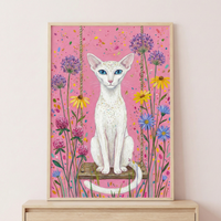 A whimsical illustration features a slender white cat with vivid blue eyes and large ears, sitting on a wooden swing. The cat has fine gold speckles on its forehead and chest. It sits poised, its long tail curling in front. The background is a vibrant pink with colorful splatters. Surrounding the cat are various stylized flowers in shades of purple, yellow, pink, and blue. The artwork is framed in a light wood frame, resting on a wooden surface against a soft pink wall.