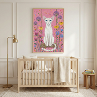 A bright nursery features a light-toned wooden crib centered in a room with neutral walls and classic molding. Above the crib hangs a large framed illustration of a slender white cat with blue eyes sitting on a swing, surrounded by tall purple and yellow flowers on a pink speckled background. A cream-colored knitted blanket is draped over the crib's side. To the left is a tall gold floor lamp, and to the right, a small wooden side table holds a stack of books on a textured rug.
