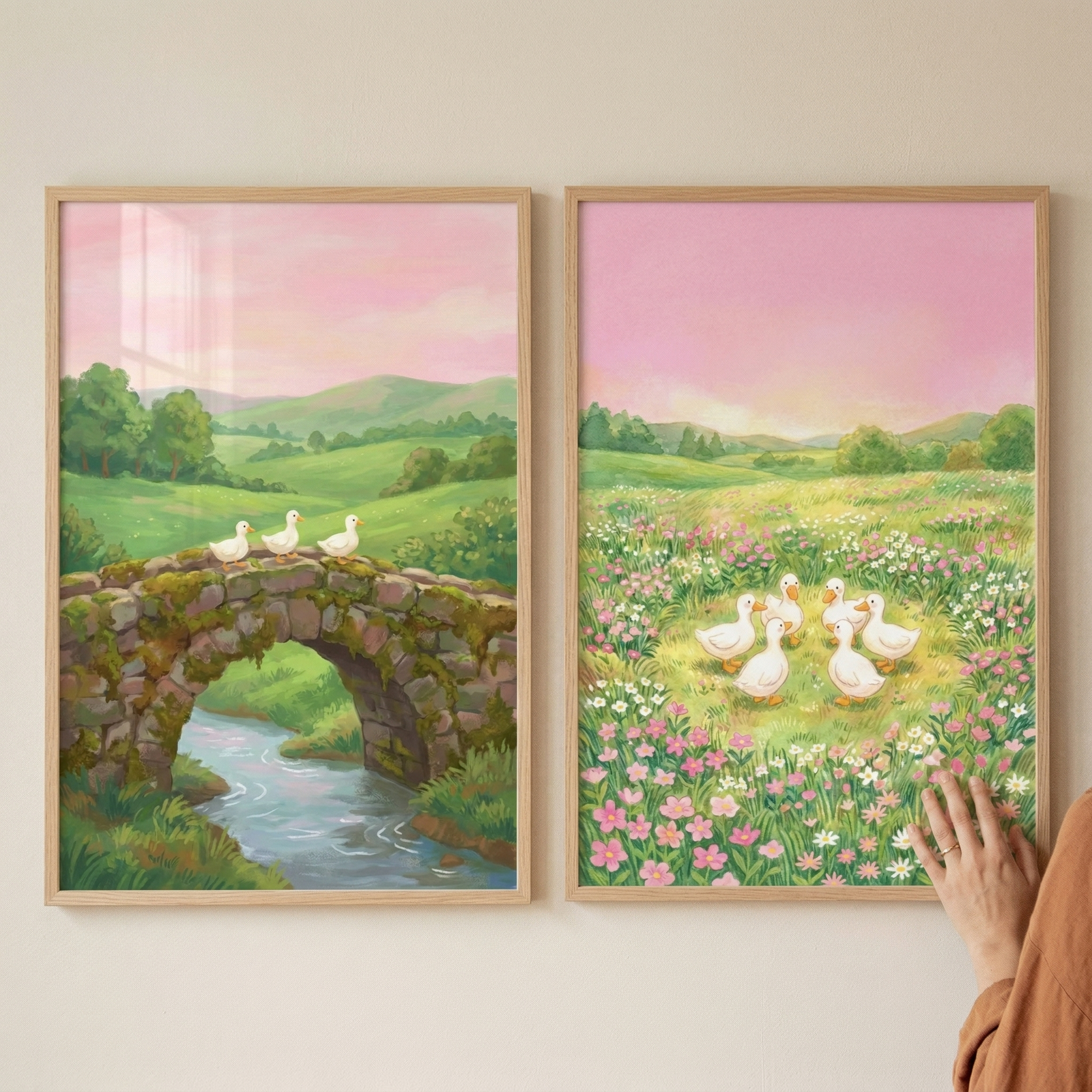 A pair of framed illustrations depicts serene, pastoral landscapes in a soft, whimsical style.

The illustration on the left features three small white ducks walking across an old, moss-covered stone bridge. Beneath the bridge, a clear blue stream flows gently through a lush green field. In the background, rolling green hills stretch toward a soft pink and yellow evening sky.

