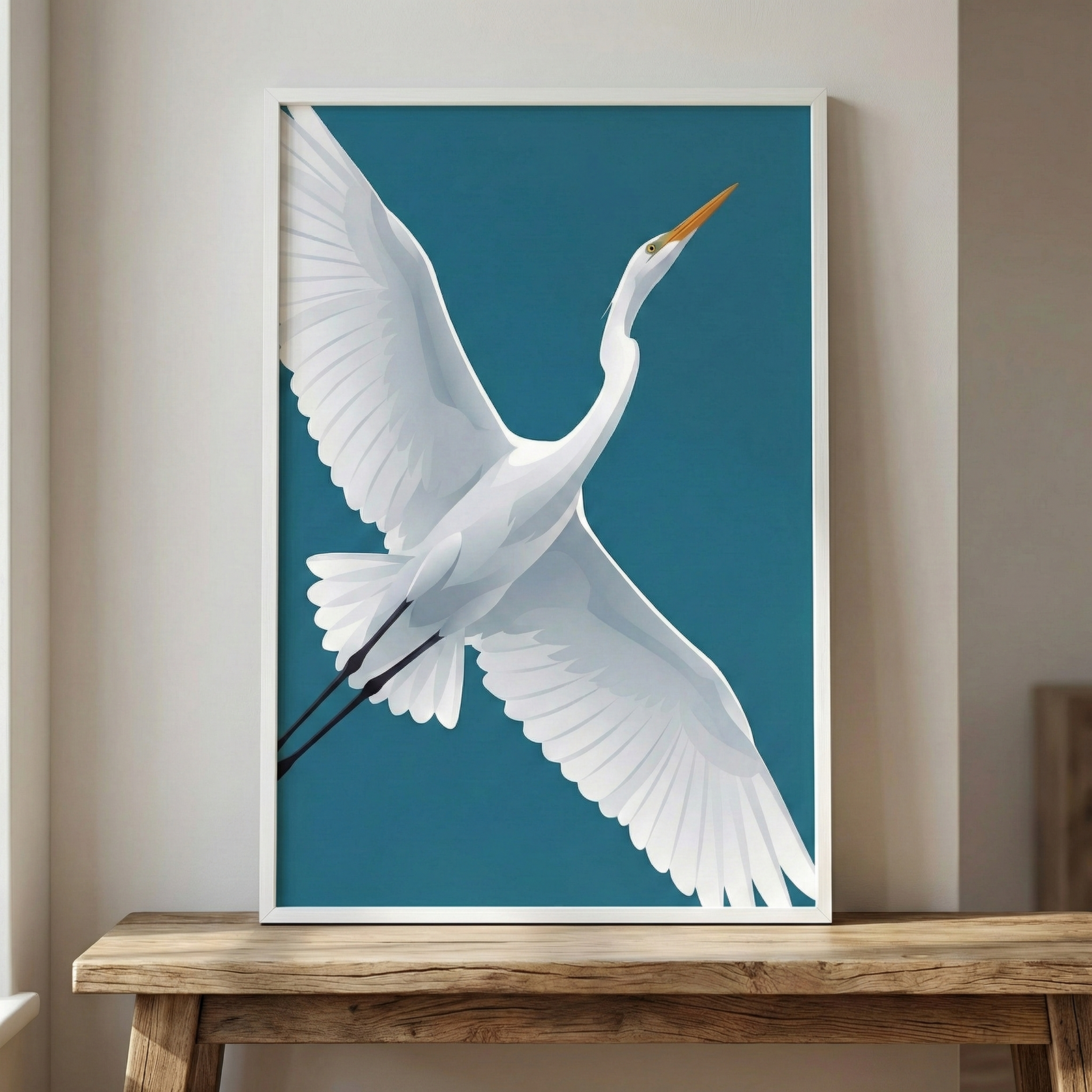 A minimalist artwork depicts a white egret in mid-flight against a solid, deep teal background. The bird is shown with its large wings fully extended, a long neck curved upwards, and a slender yellow beak. Its thin black legs are tucked behind as it soars. The piece is presented in a clean white frame and is displayed on a rustic wooden table against a neutral, light-colored wall.