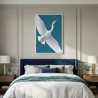 A vertical framed artwork depicting a white egret in flight against a teal background is centered above a bed. The bed features a plush navy blue velvet headboard and is styled with white linens, a teal throw blanket, and a variety of pillows. The light taupe wall is decorated with classic panel molding. On each side of the bed, wooden nightstands hold gold-based lamps with white shades. The scene is elegant and calm, with soft natural light highlighting the coordinated decor.