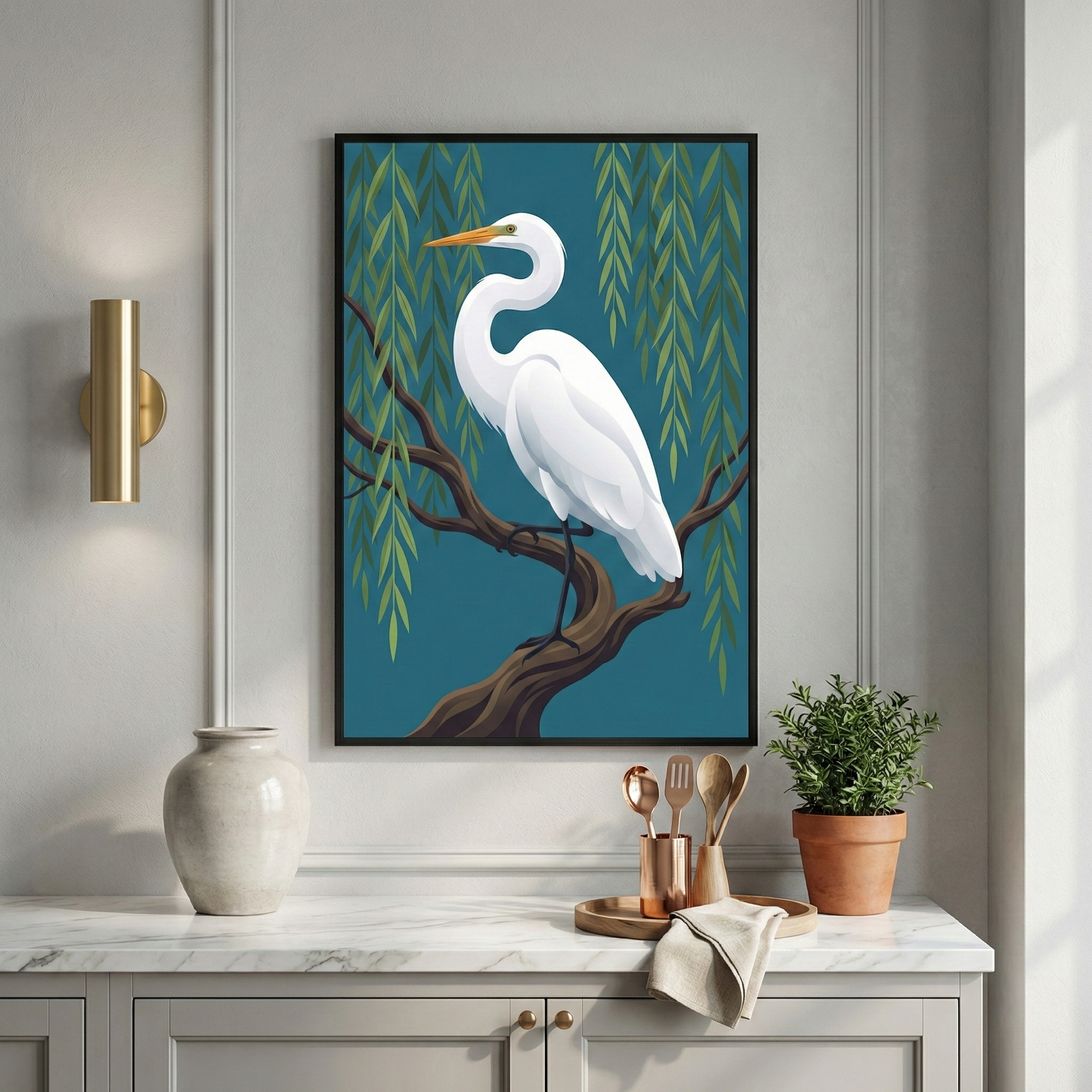 A framed piece of artwork hangs on a light gray wall above a marble countertop. The illustration features a large, elegant white bird with a long neck and a yellow beak, perched on a dark brown branch. Behind the bird, long, slender green leaves hang from willow branches against a deep teal background. To the left is a gold-toned wall light. Below, on the counter, are a large white vase, a tray with wooden utensils in a copper cup, and a small potted plant in a clay pot.