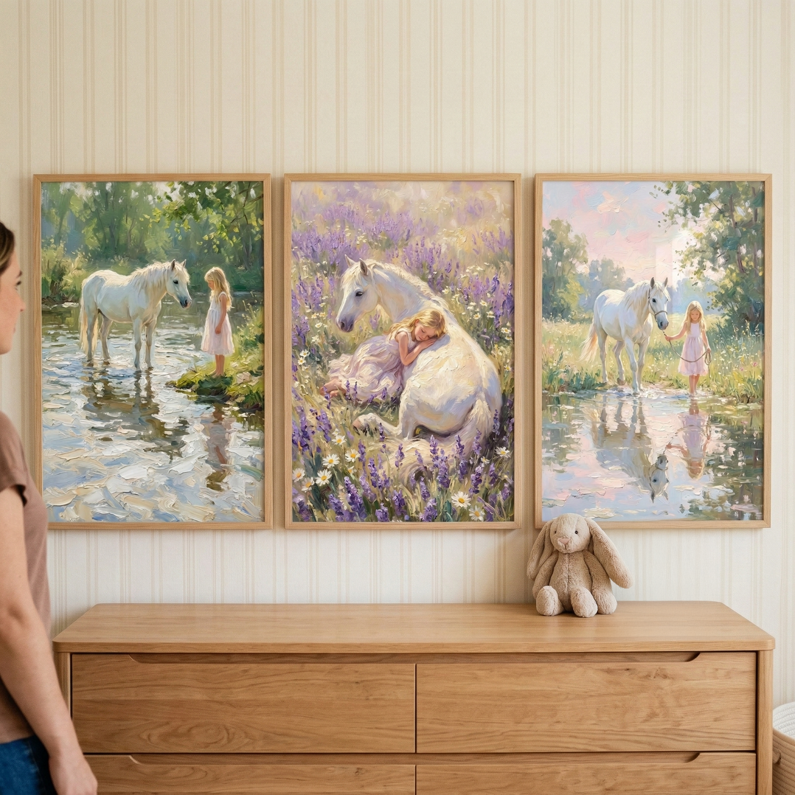 Three thin white frames rest on a rustic wooden table against a neutral wall, with a person's hand visible as they adjust the rightmost frame. Each frame showcases an art piece featuring a white horse and a young girl in a pink dress in a different natural setting. The left artwork depicts the pair by a calm stream. The center piece shows them resting together in a field of purple wildflowers. The rightmost illustration shows the girl leading the horse through shallow water. 