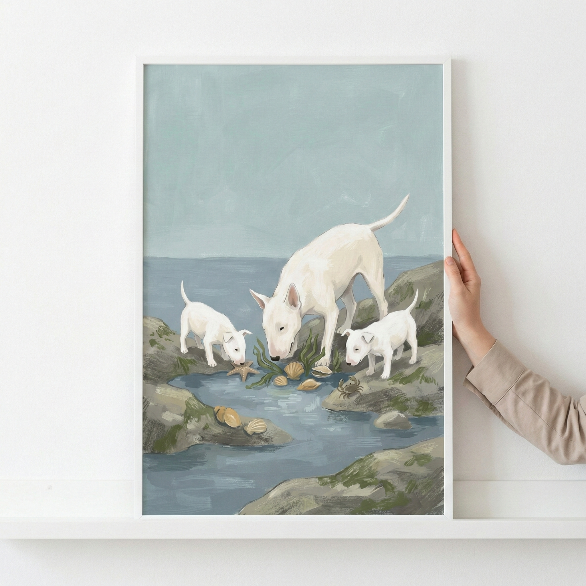 An illustration depicts three white dogs with distinctive egg-shaped heads—an adult and two puppies—at the edge of a rocky tide pool. They are looking down at seashells and a starfish scattered among the rocks and shallow water. The scene is rendered in a soft, textured style with a color palette of pale blue, seafoam green, and earthy grays. The piece is encased in a white frame, which is being held against a white wall.