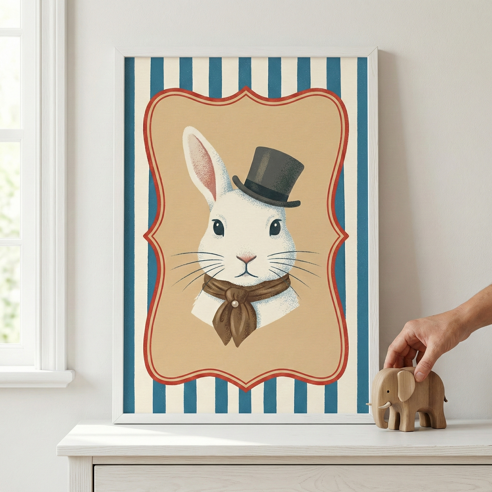 A framed vertical illustration of a white rabbit wearing a black top hat and a brown necktie sits on a white wooden dresser. The rabbit is centered inside a tan, decorative scalloped frame with a red border, set against a background of vertical blue and white stripes. To the left of the white frame, a portion of a white-paned window is visible. To the right, a person's hand touches a small, carved wooden elephant figure resting on the dresser.