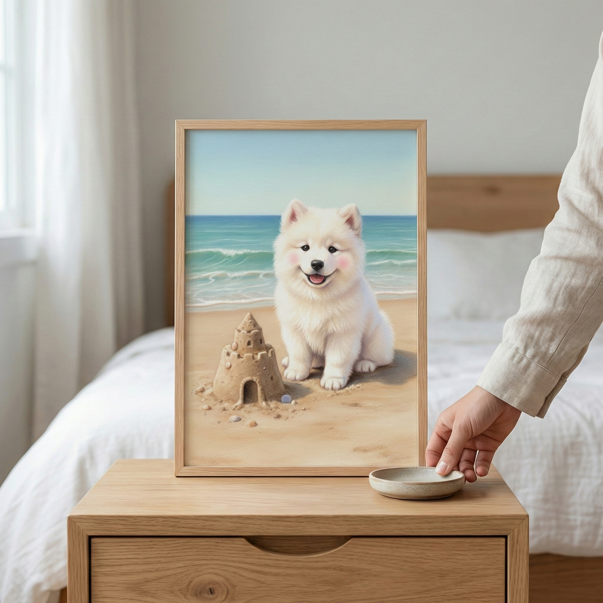 A framed illustration of a fluffy white puppy with pink cheeks sits on a light wooden nightstand. In the artwork, the puppy smiles next to a detailed sandcastle on a sunny beach, with gentle ocean waves in the background. The frame is positioned in a minimalist bedroom next to a bed with white linens. A person's hand reaches for a small ceramic dish on the table. The entire scene is bathed in soft, natural light, creating a serene and cozy atmosphere.