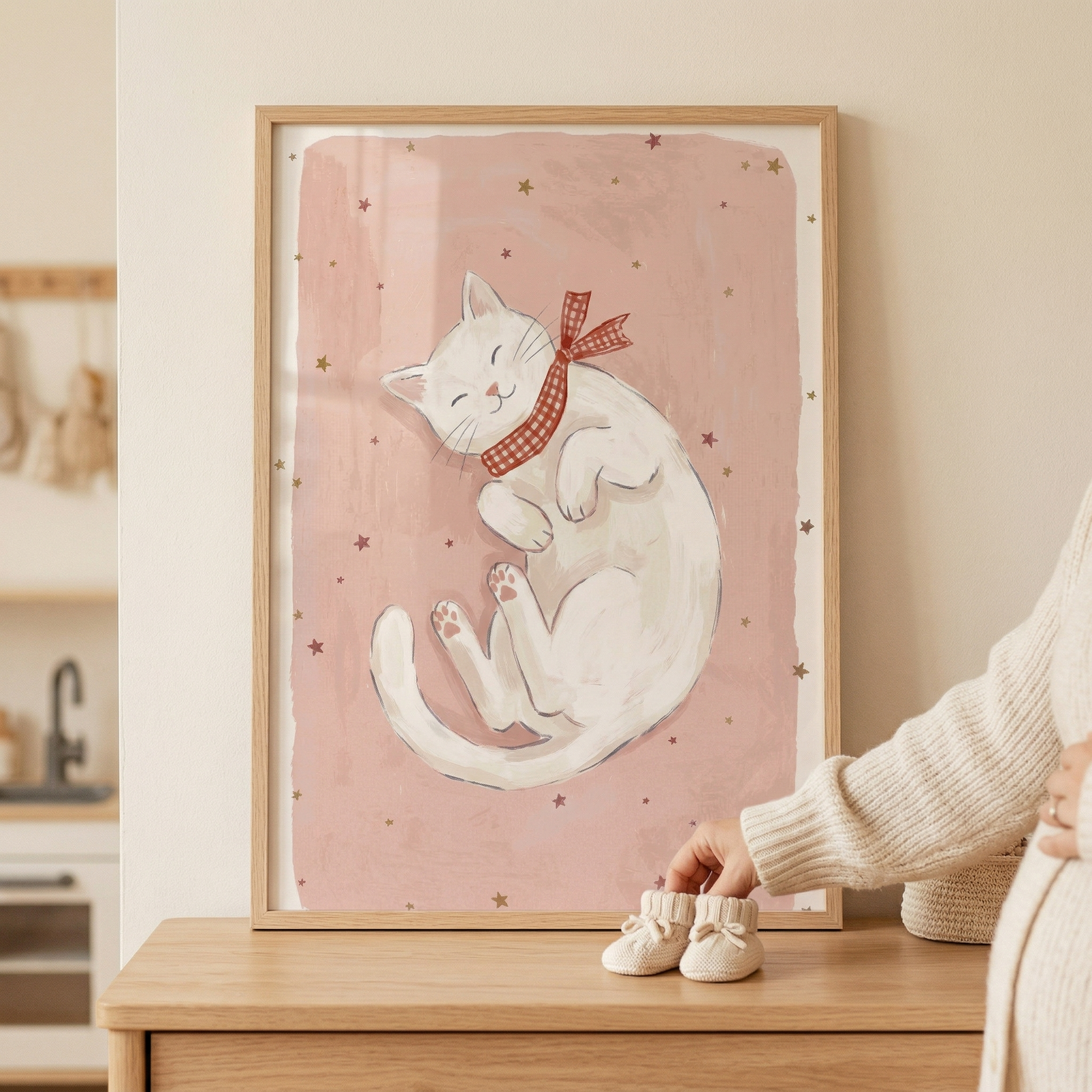 A framed piece of art rests on a light wood tabletop, showcasing a cute white cat curled up on its back against a soft pink background dotted with small stars. The cat wears a red and white checkered bow. In front of the frame sit a pair of small, cream-colored knitted baby booties, with a hand reaching toward them. A person wearing a cream-colored knit sweater is partially visible on the right. The overall scene has a warm, gentle aesthetic.