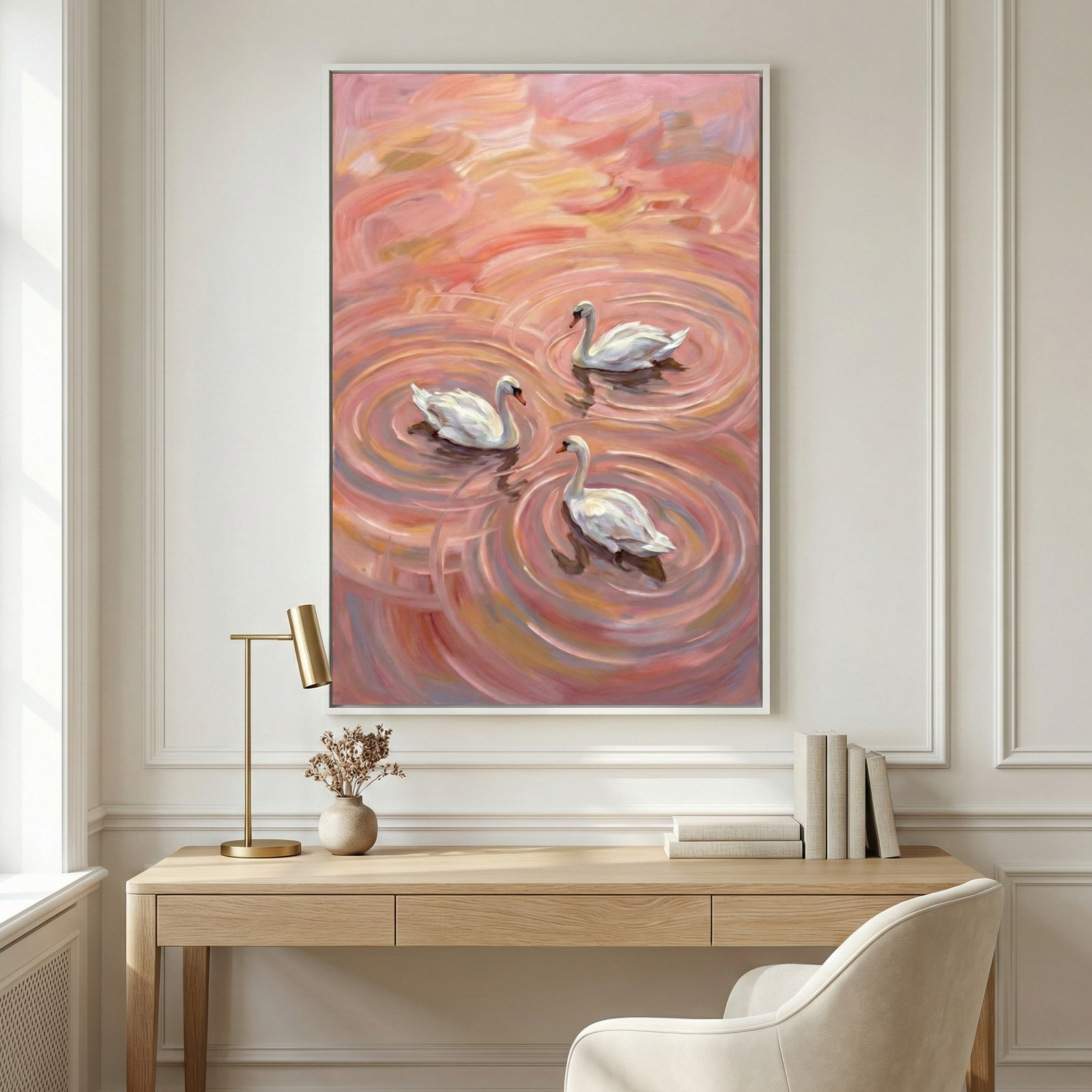 An interior view of a serene room featuring a large framed vertical canvas hanging on a paneled wall. The artwork depicts three elegant white swans floating in a sea of vibrant pink, rose, and peach-colored ripples, created with bold, expressive brushwork. Below the wall decor is a light wood desk with a clean design. The desk's surface holds a slender brass lamp on the left, a small neutral-toned vase with delicate dried sprigs, and several books in white and gray covers arranged neatly on the right. 