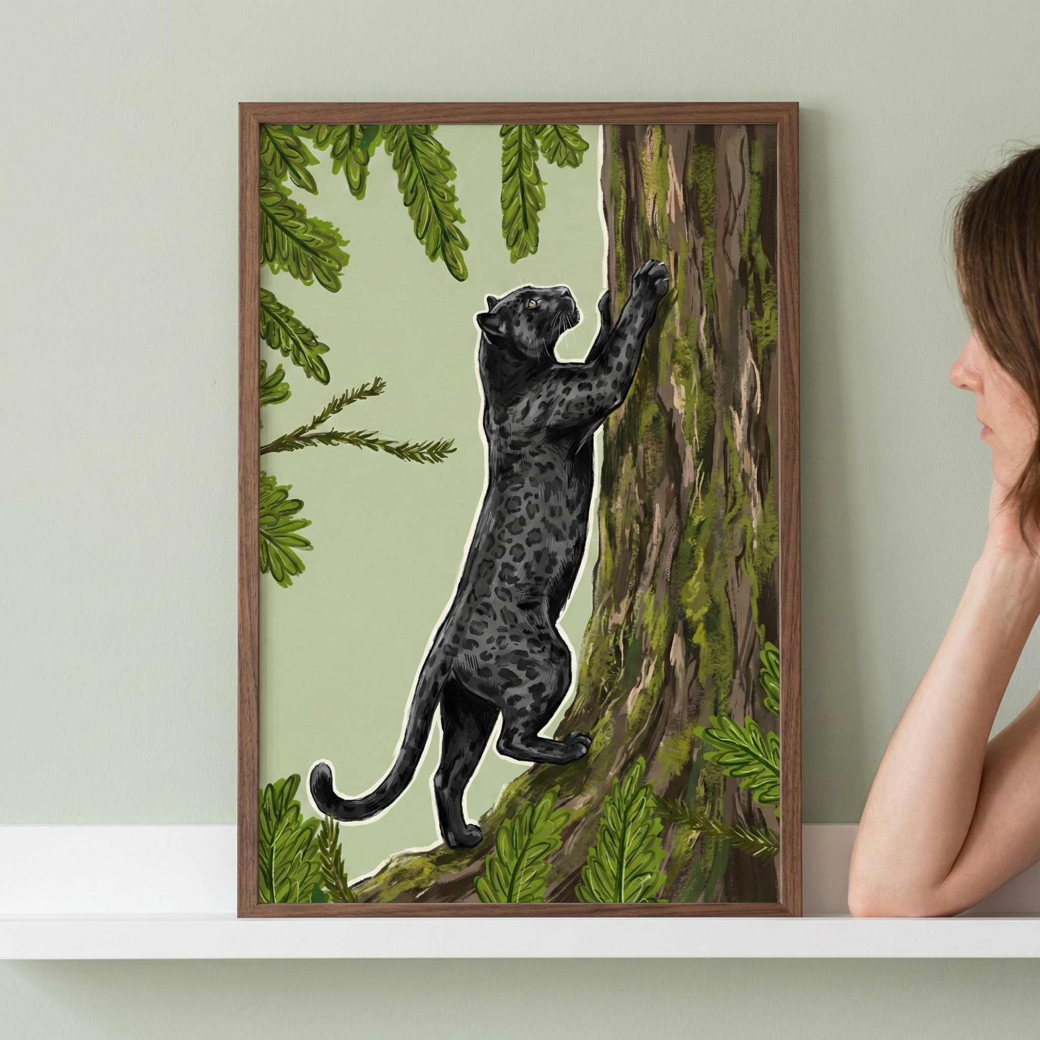 A framed illustration depicts a black leopard with faint rosettes climbing a mossy tree trunk. The big cat reaches upwards, its dark body contrasted against a pale green background and framed by lush green foliage. The art is in a simple brown wooden frame, resting on a white shelf. On the right, a person's profile and arm are partially visible as they look at the piece.