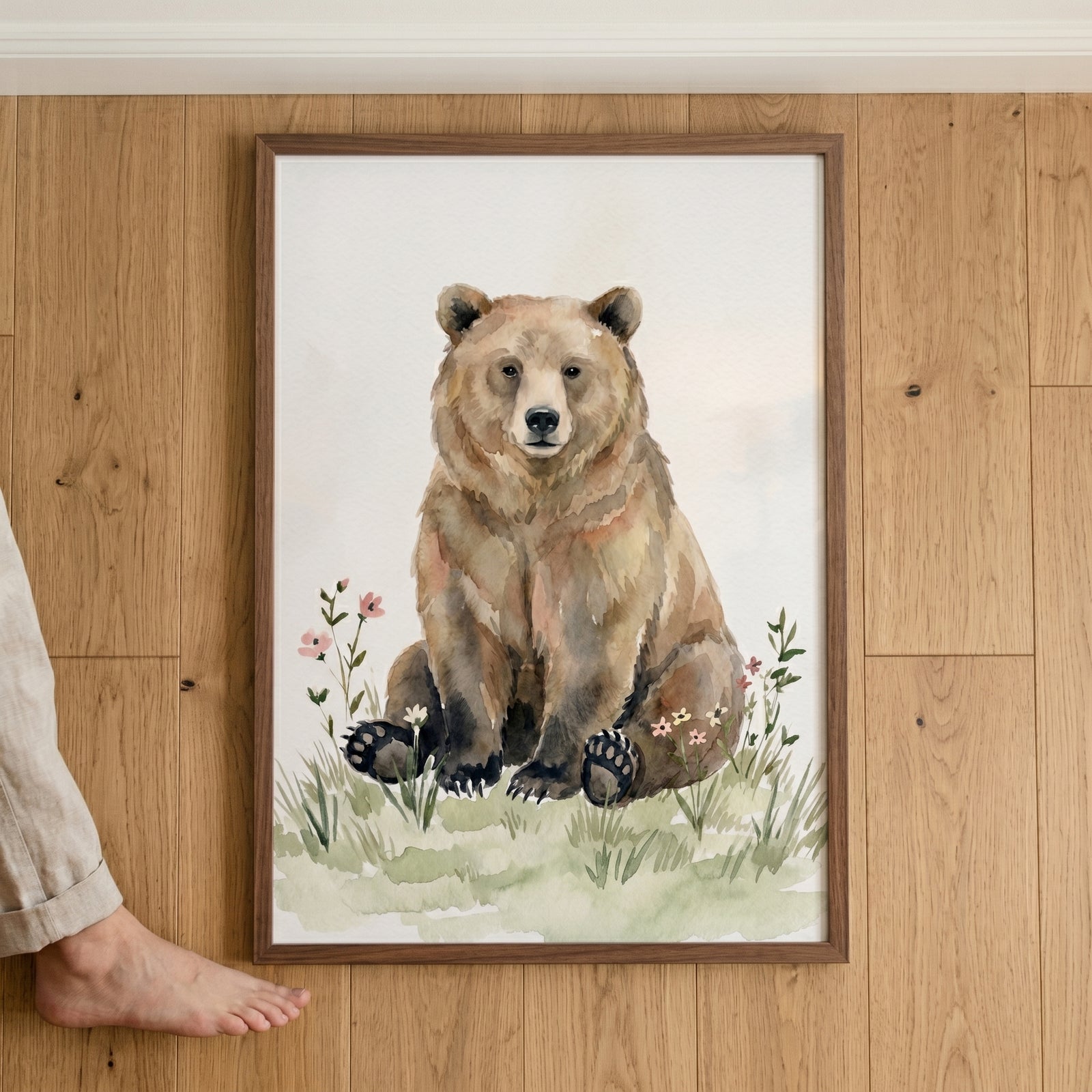 Framed wildflower brown bear wall art leaning on a wood floor, featuring a bear sitting in a field with pink flowers.