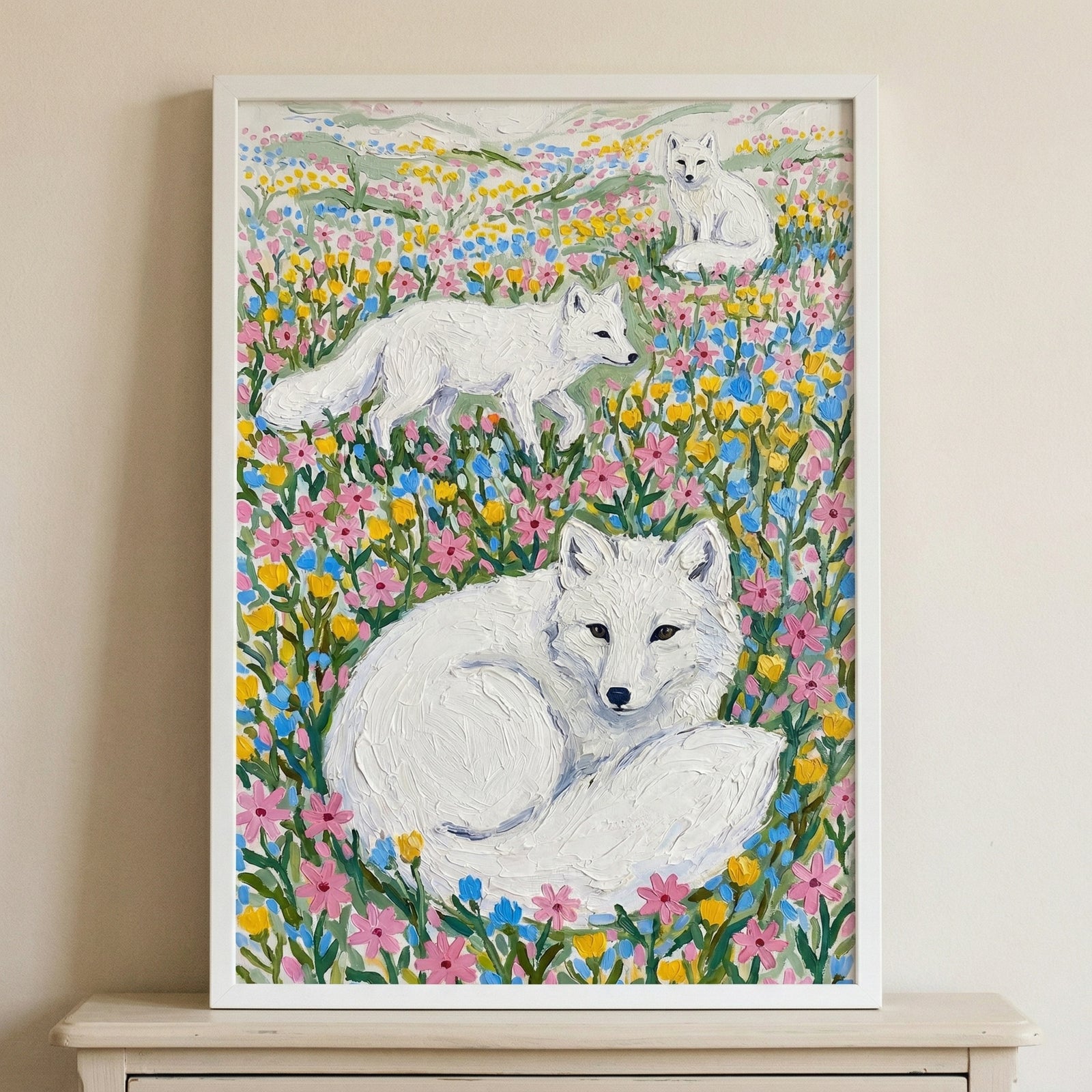 Framed painting of three white foxes resting and walking through a dense field of pink yellow and blue wildflowers fills the canvas with soft texture and color. The artwork leans against a neutral wall creating a gentle whimsical nature inspired focal point.