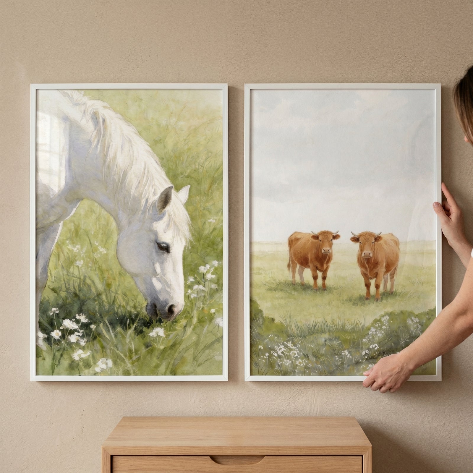 Hands adjust a wildflower meadow farm animal wall art set of 2 featuring a grazing white horse and two brown cows.