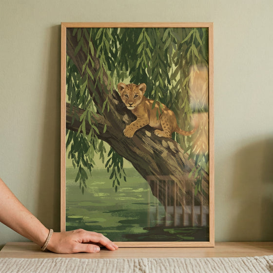 A framed illustration featuring a young lion cub perched on a thick tree branch. The cub, with its spotted coat and curious expression, is surrounded by the long, drooping green leaves of a tree that hangs over a tranquil body of water. The artwork uses a soft, natural color palette of greens, browns, and earthy tones, giving it a peaceful, nature-themed feel. The entire piece is enclosed in a simple, light-colored wooden frame. In the foreground, a person's hand and wrist with a woven bracelet rest on a wo